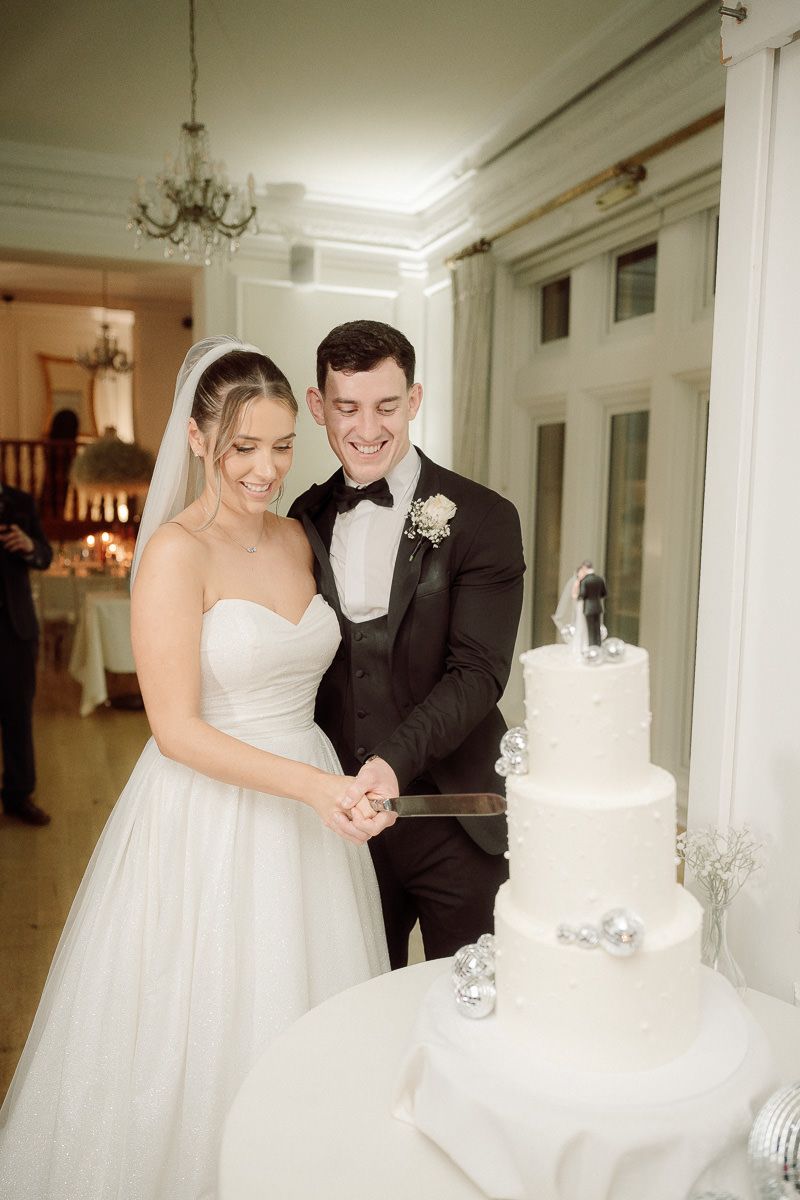 A couple in formal wedding attire shares a sweet moment while cutting their elegant white tiered wedding cake.