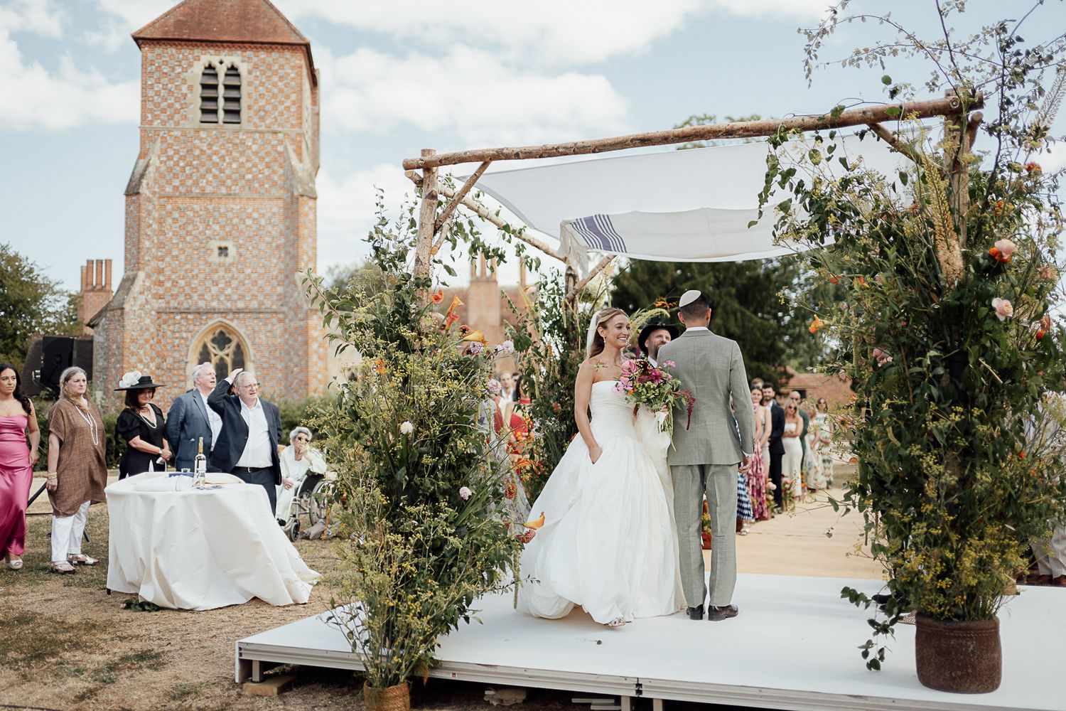 Outdoor wedding ceremony on a raised platform with floral arch, bride and groom, and a historic brick church tower backdrop.