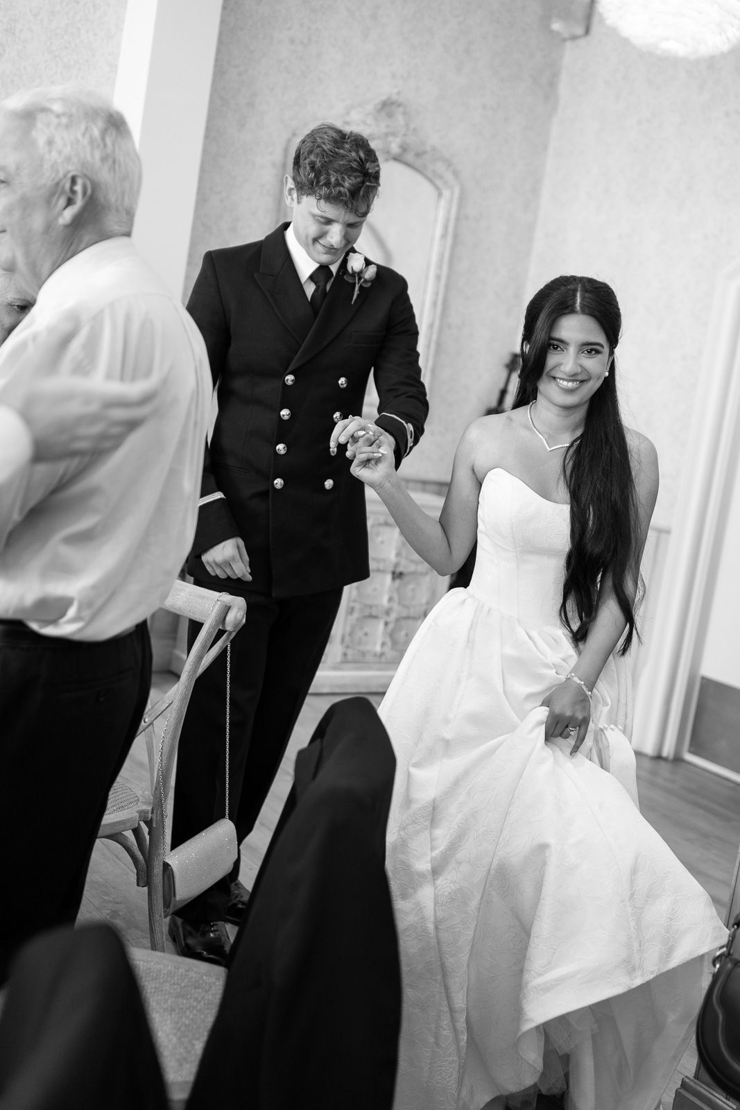 Black and white photo of smiling bride and groom in naval suit walking hand in hand at wedding ceremony.