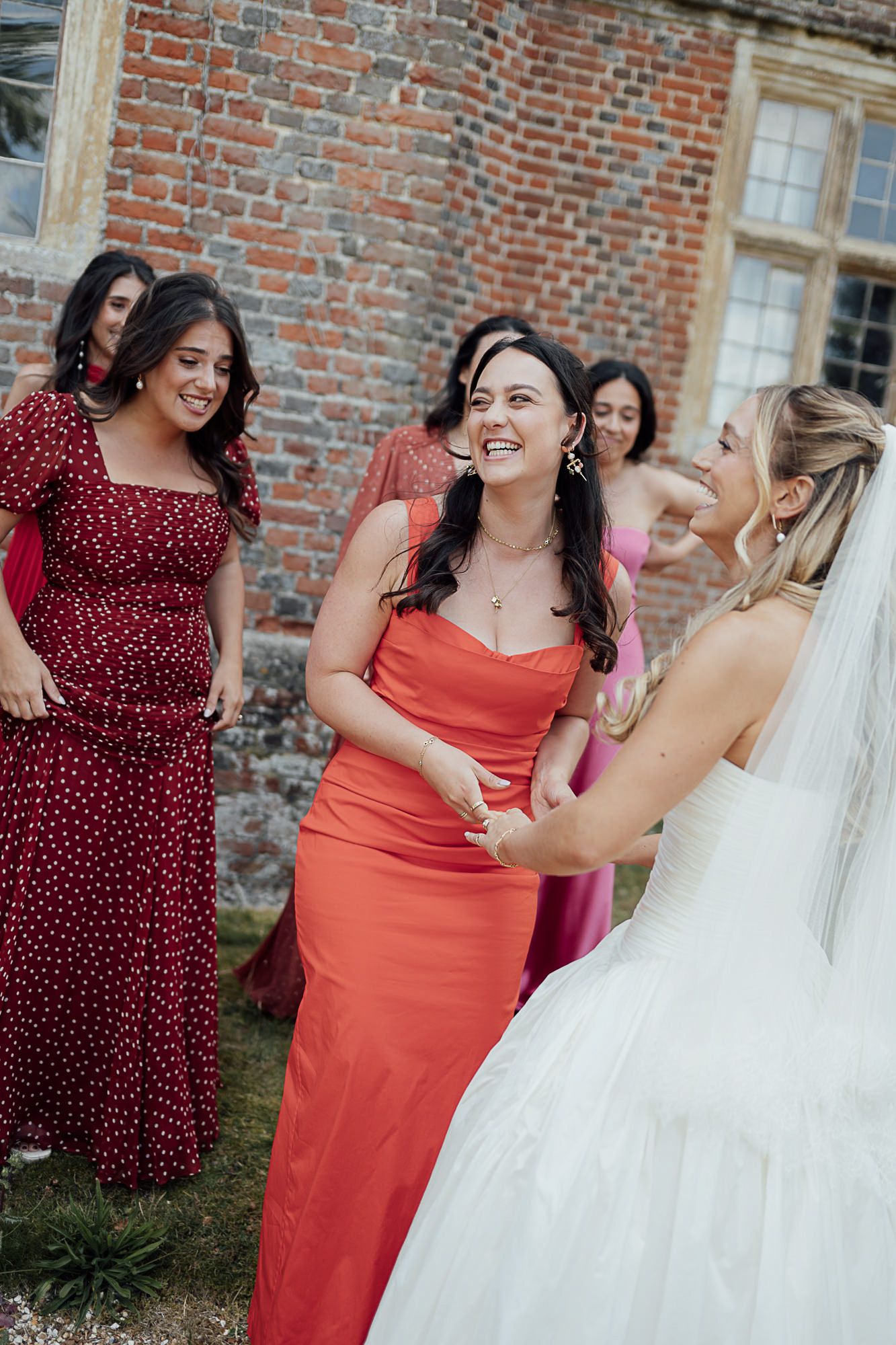 Bridesmaids in red and pink dresses laughing with a bride in a white gown outside a brick venue.