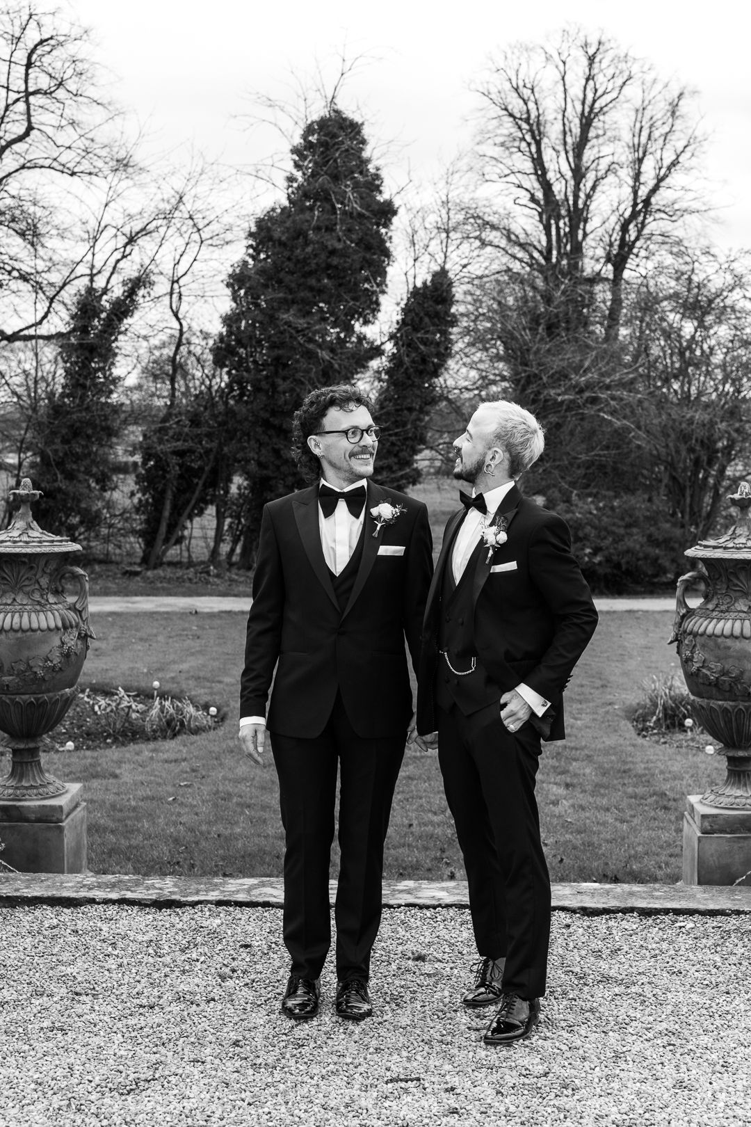 Two elegantly dressed people in tuxedos share a moment on a gravel path with ornate garden urns in black and white.