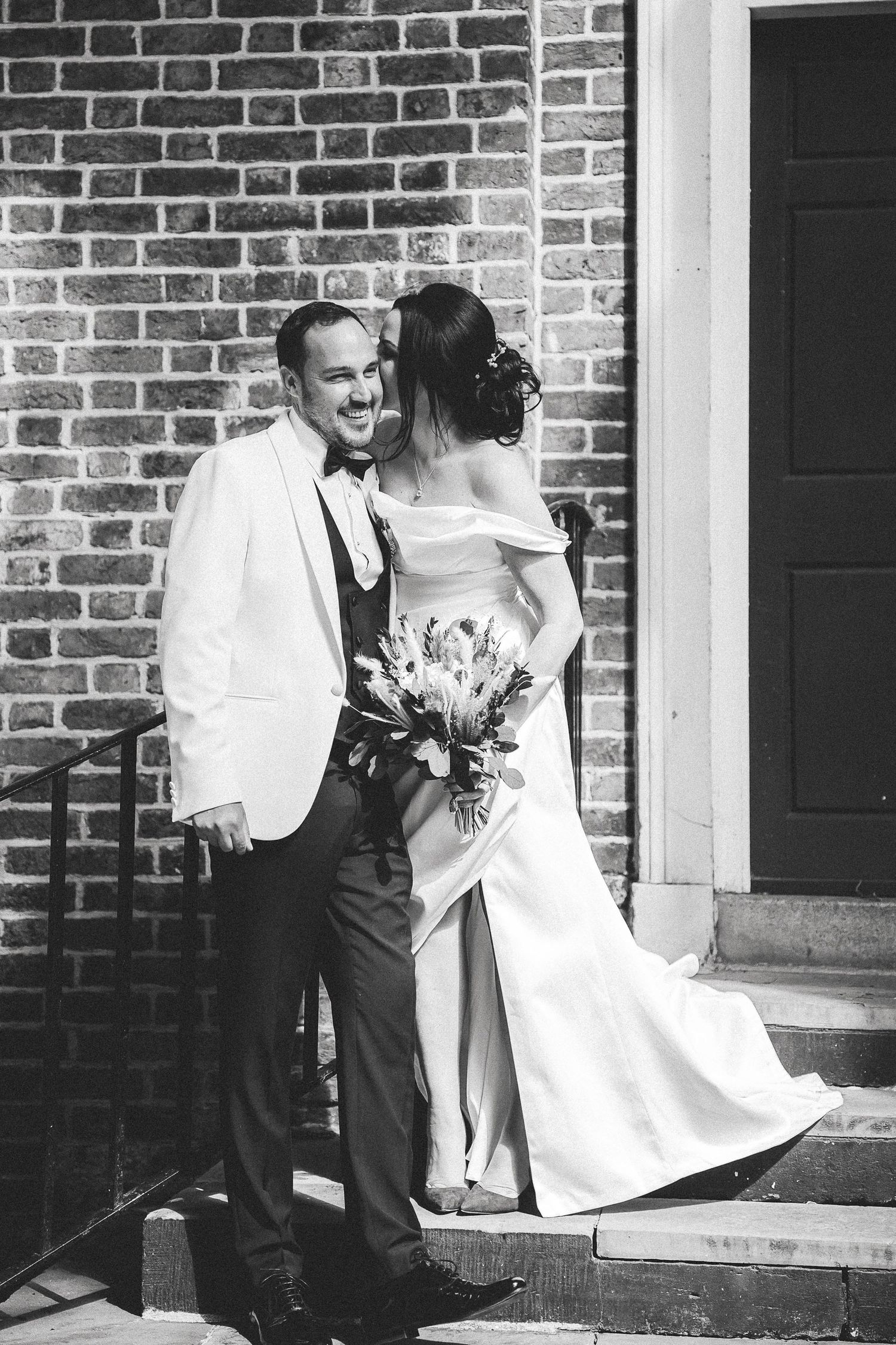 A bride in a white dress descends stairs with help from someone wearing a white jacket and dark pants by a brick wall.
