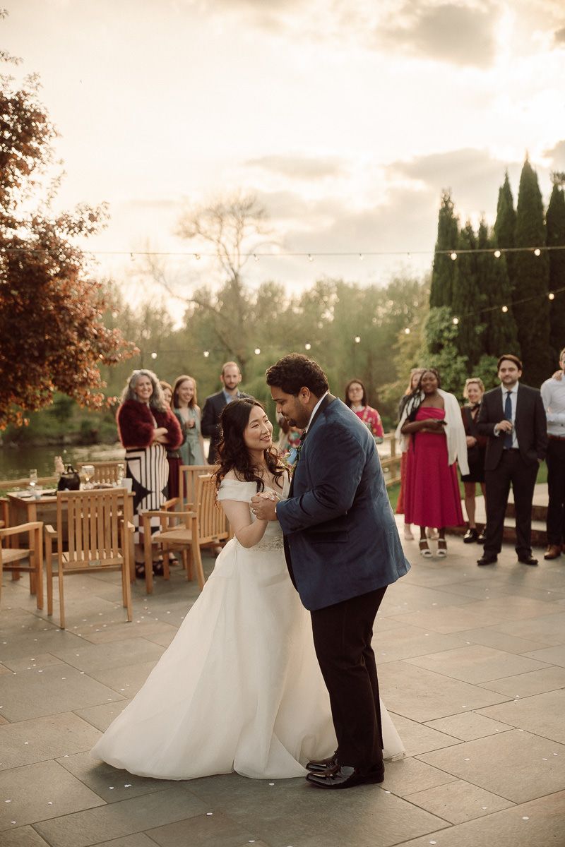 A couple shares their first dance on an outdoor patio while wedding guests look on during a sunset reception.