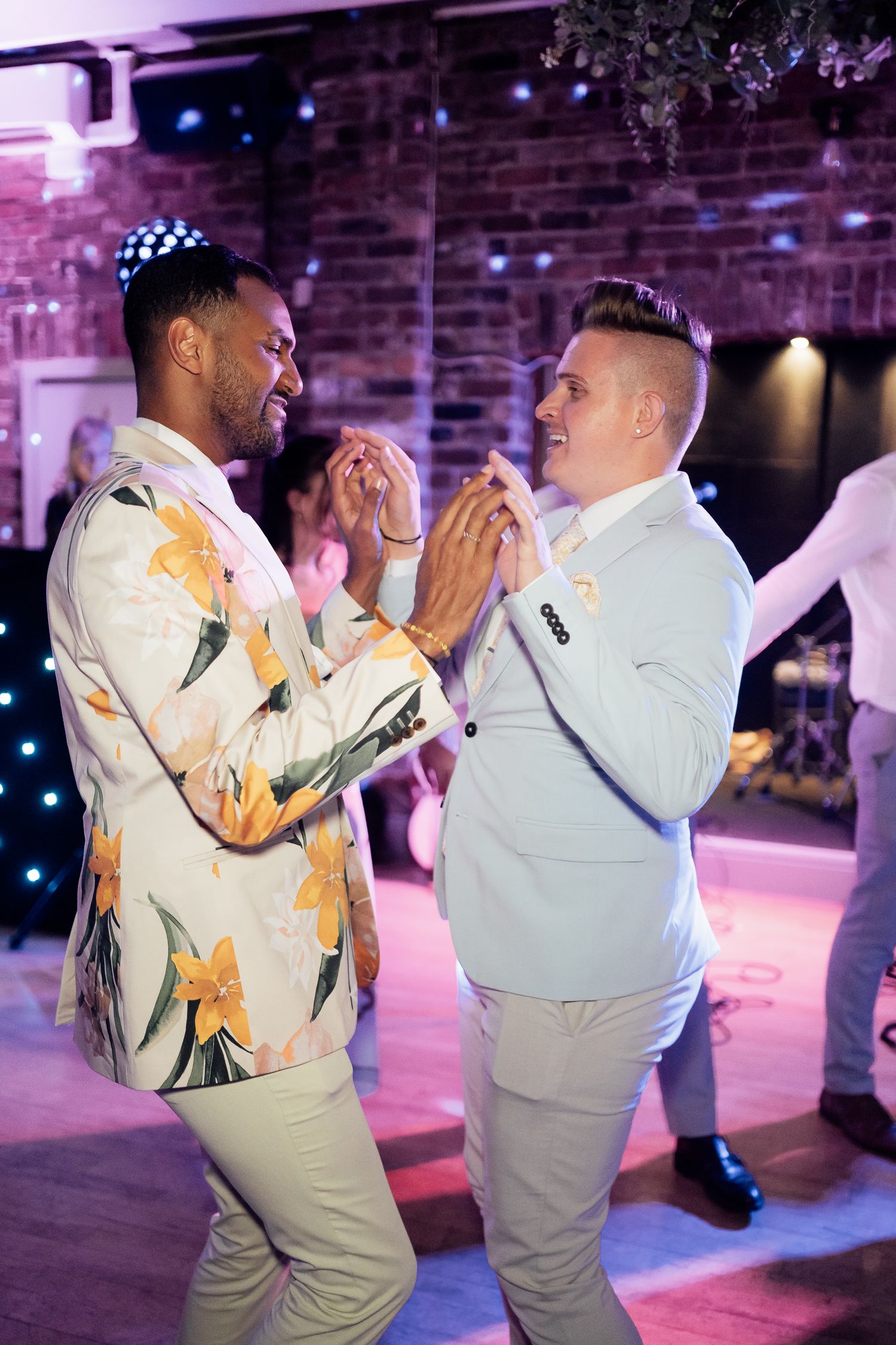 Two people in light colored suits dance together at a party with purple lighting and exposed brick walls in the background.