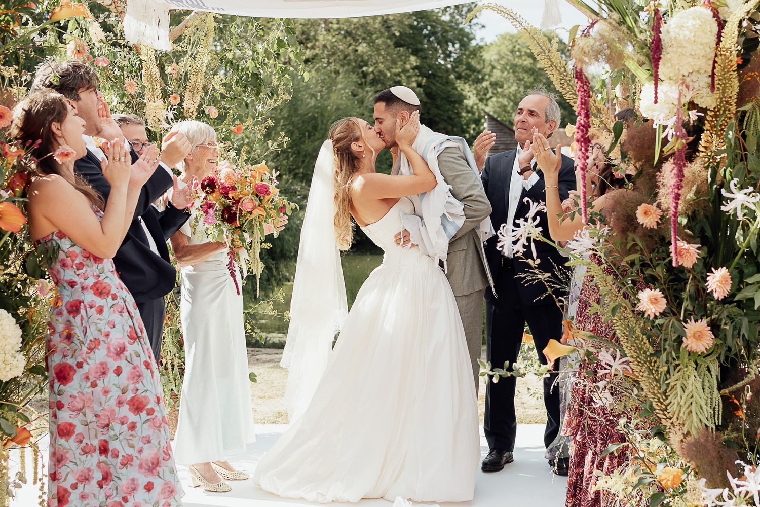 Bride and groom share a kiss at an outdoor wedding ceremony surrounded by colorful floral arch and guests.