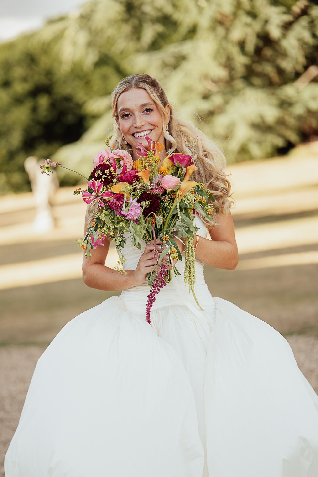 Smiling bride in white ball gown holding a vibrant colorful bouquet of pink, red, and yellow flowers outdoors.
