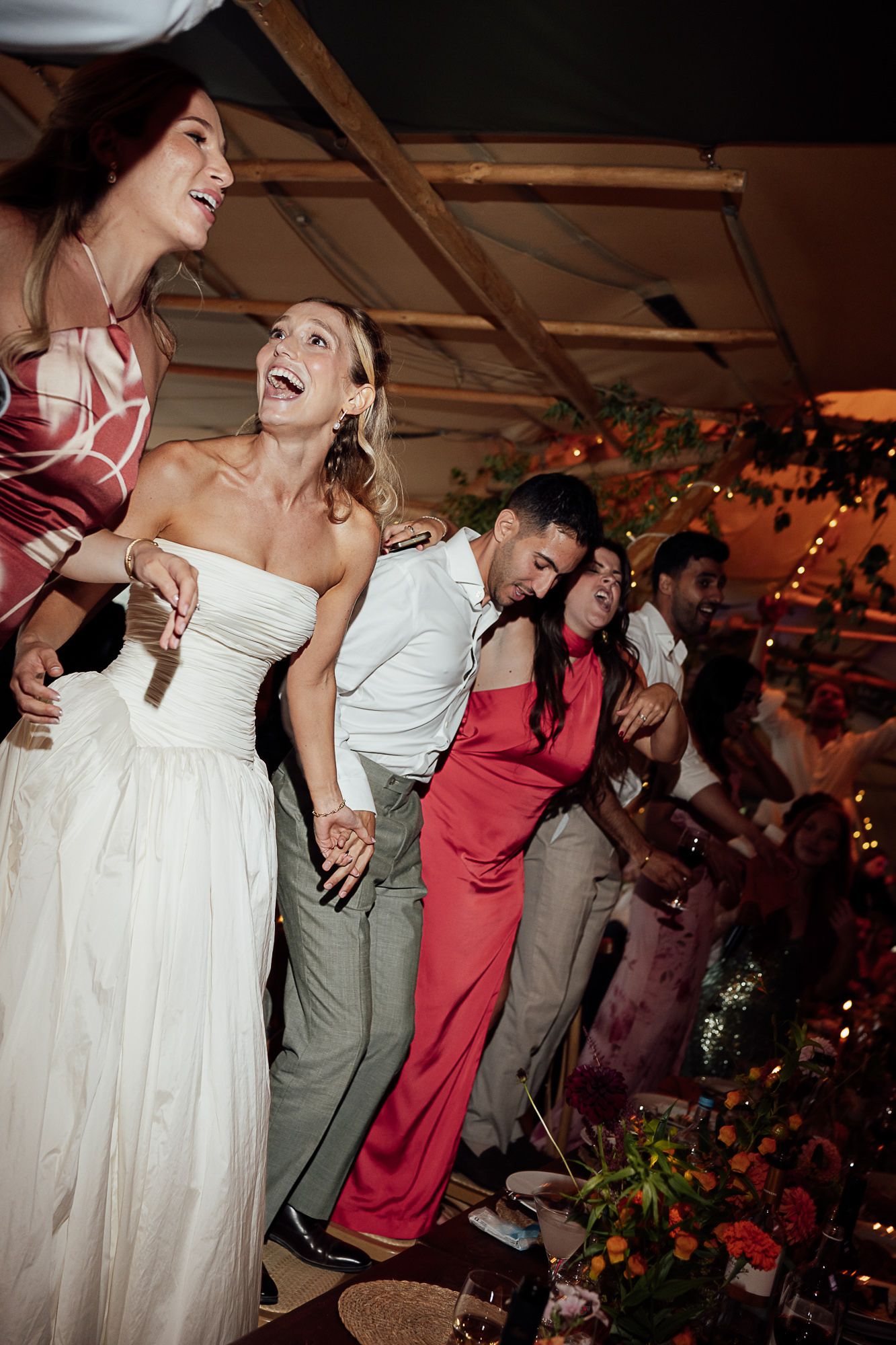 Bride in white strapless gown laughing with guests dancing joyfully at a wedding reception celebration.
