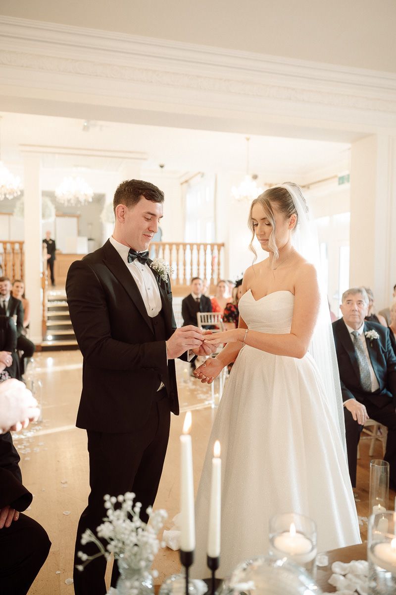 A couple exchanges rings during an elegant indoor wedding ceremony in a bright white venue.