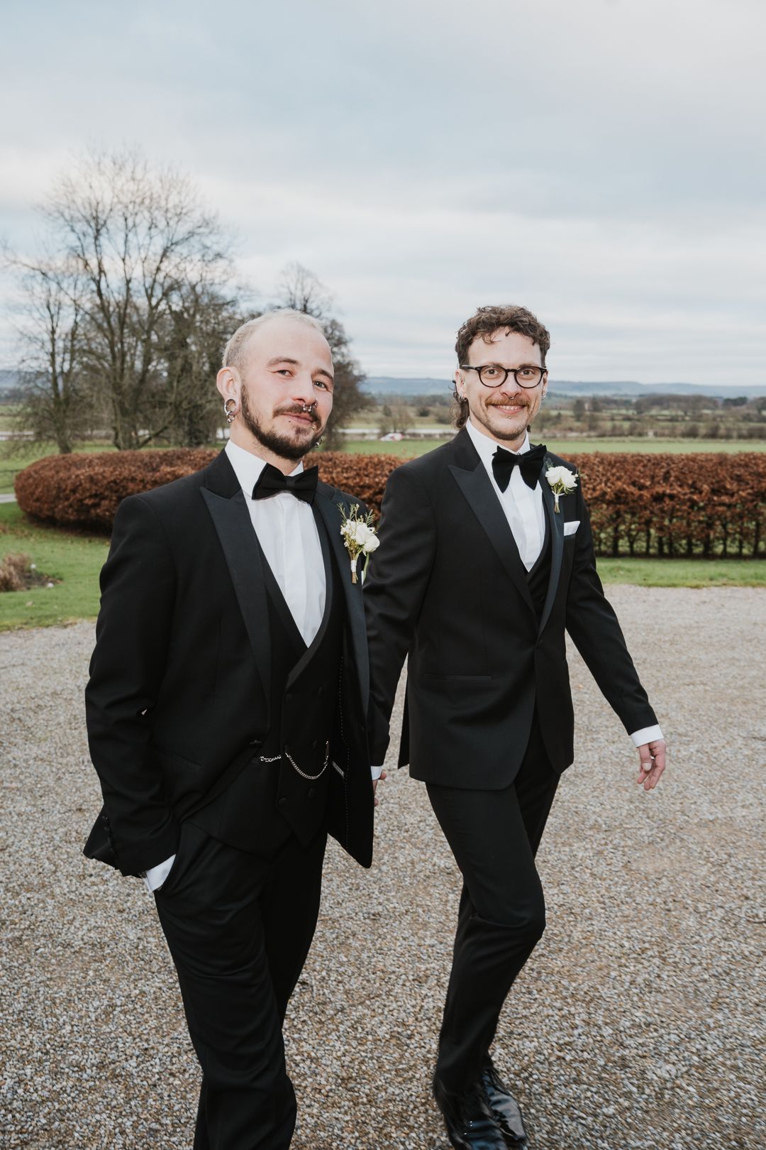 Two people in black tuxedos with bow ties walking outdoors with a scenic countryside landscape in the background.