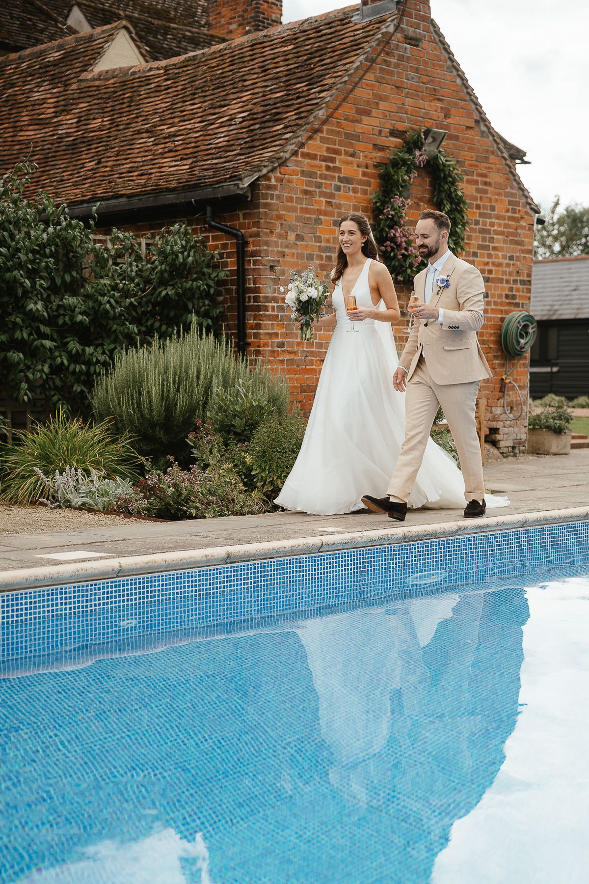 A couple sits poolside in wedding attire on a bright day next to a tiled blue swimming pool by a brick building.