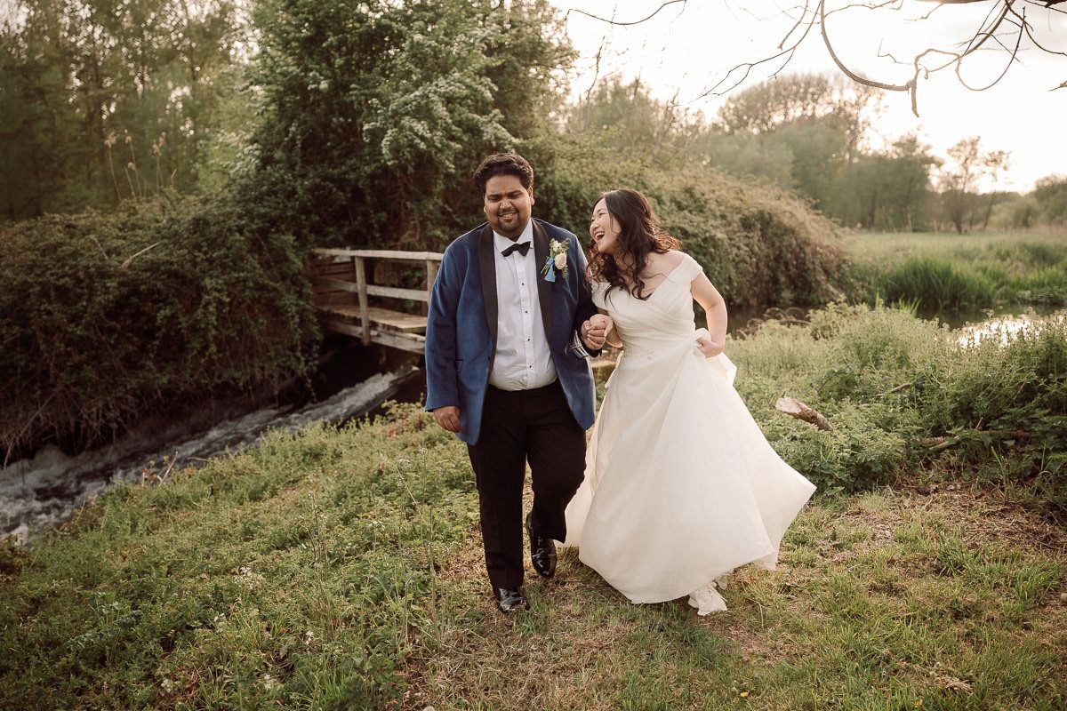 A couple in formal wedding attire walks hand in hand along a rustic path surrounded by lush greenery at sunset.