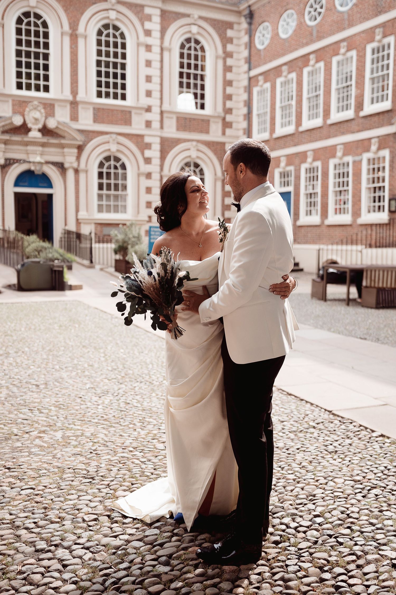 A couple shares a romantic moment in front of a historic brick building with elegant arched windows and cobblestone courtyard.