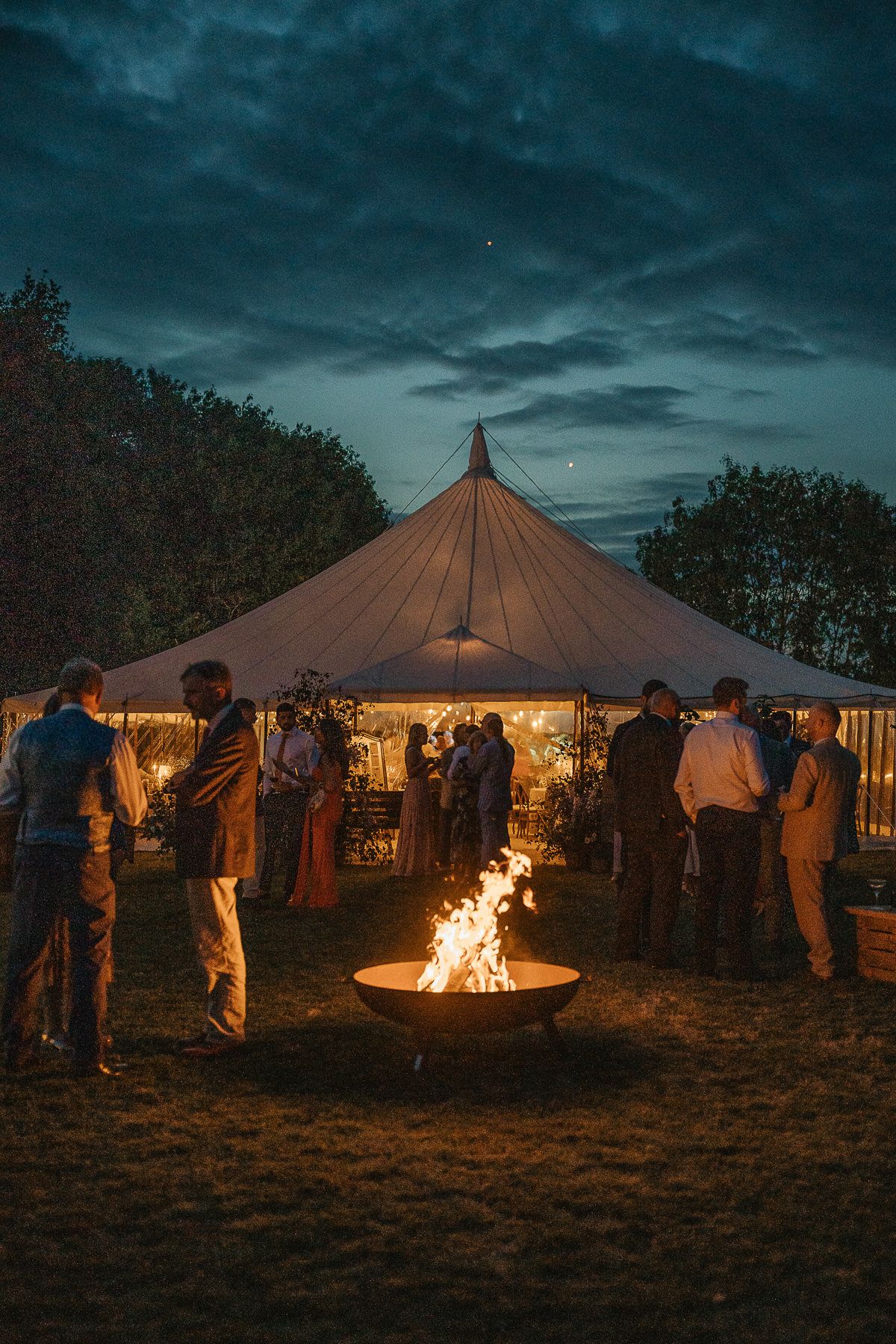 Illuminated festival tents and campfires create a cozy nighttime gathering under stormy skies.