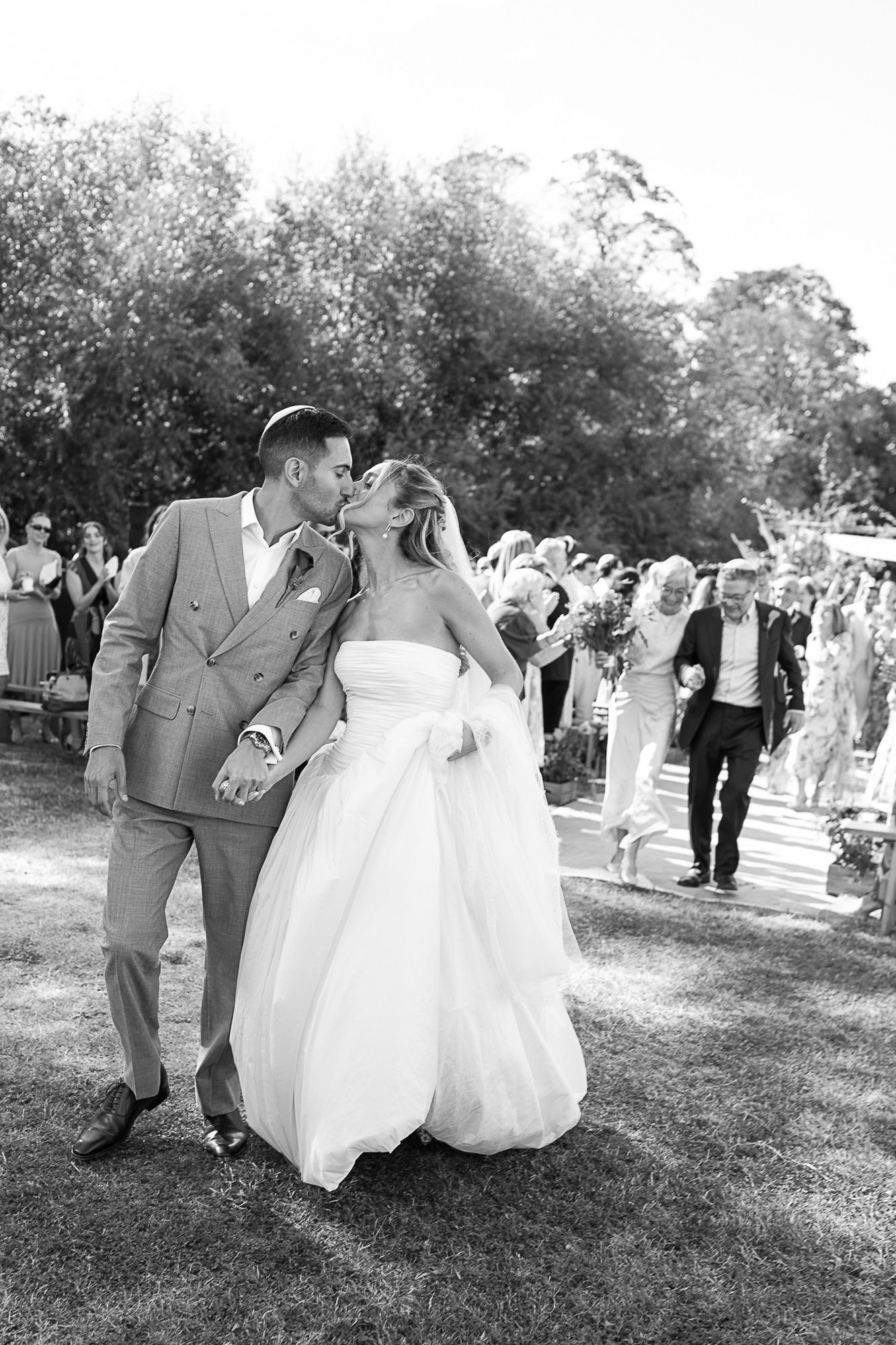 Black and white photo of a newlywed couple sharing a kiss while walking down the aisle at an outdoor wedding ceremony.