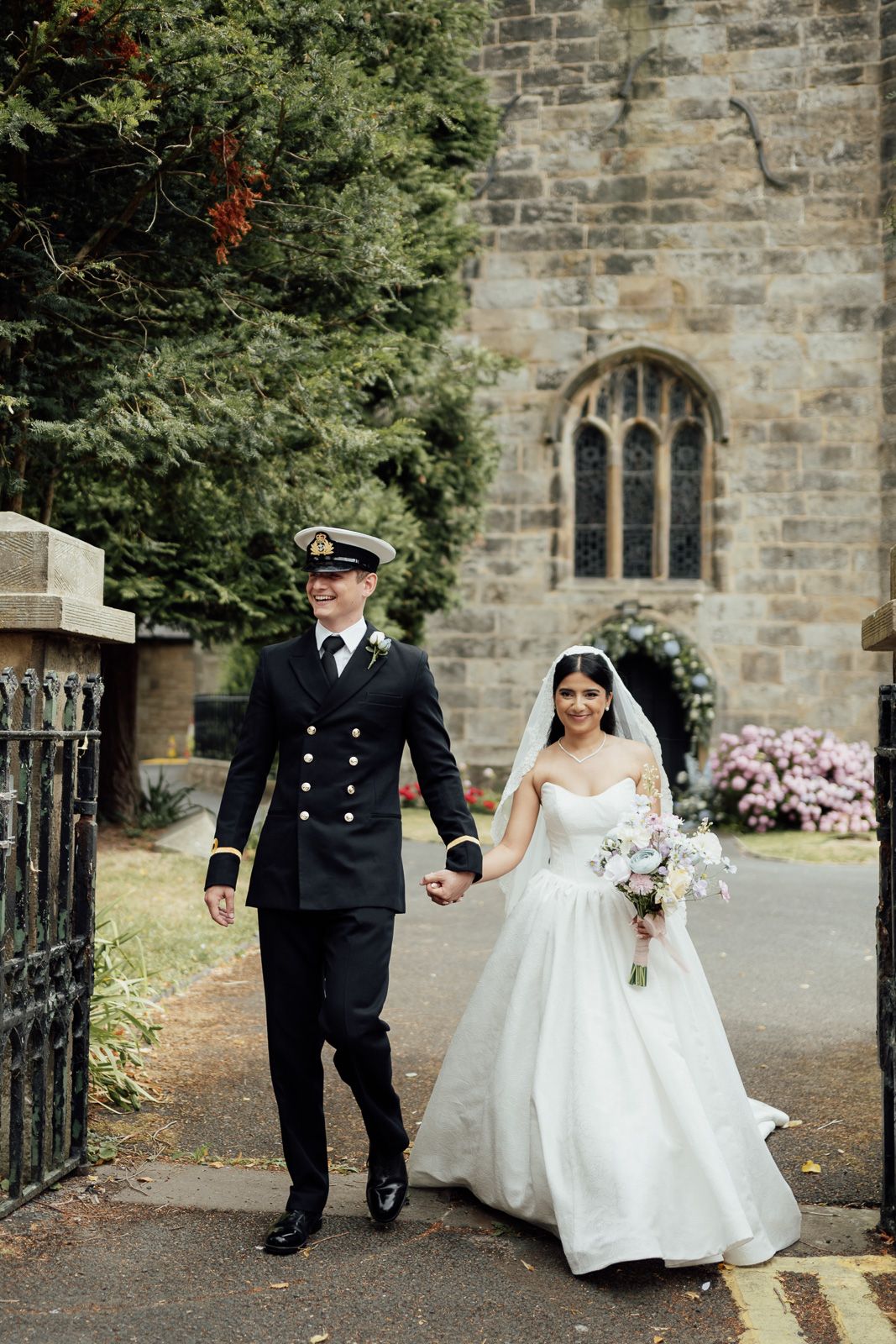 Bride in white gown and groom in naval uniform walk hand-in-hand outside a stone church.