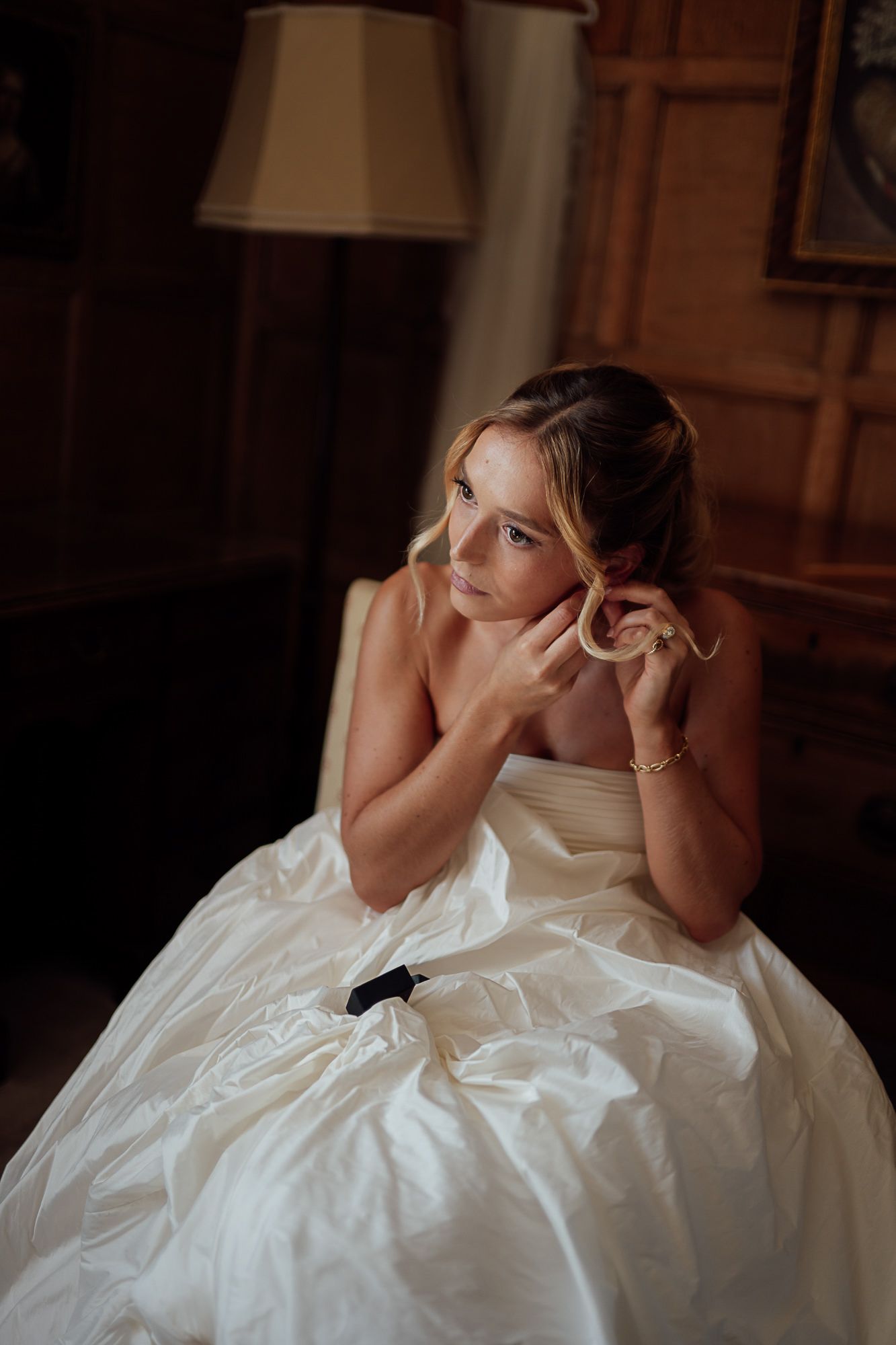 A bride in a white strapless gown sits elegantly, adjusting her earring in a warmly lit, wood-paneled room.