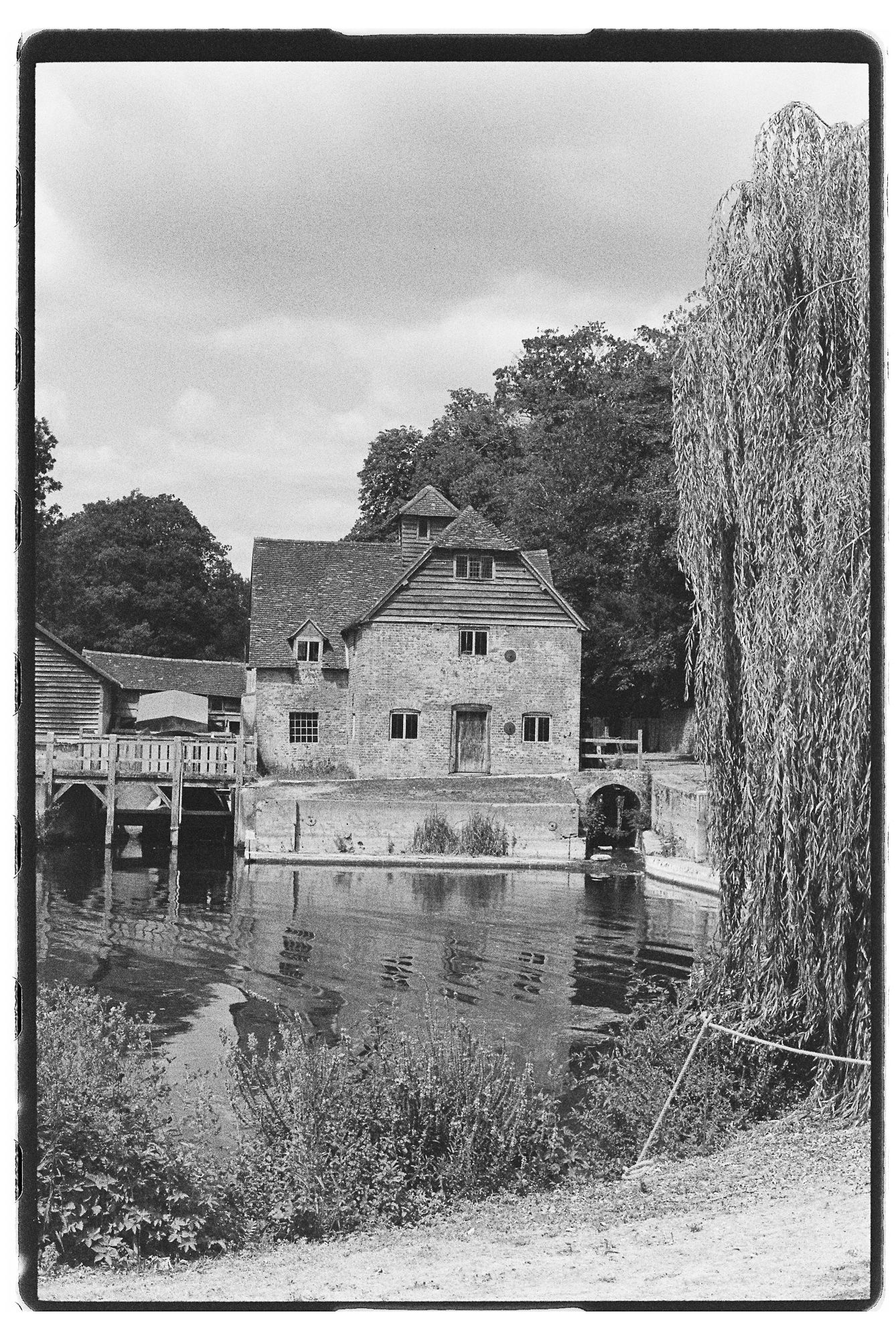 Black and white photo of a historic stone mill beside a calm reflective pond, framed by a weeping willow tree.
