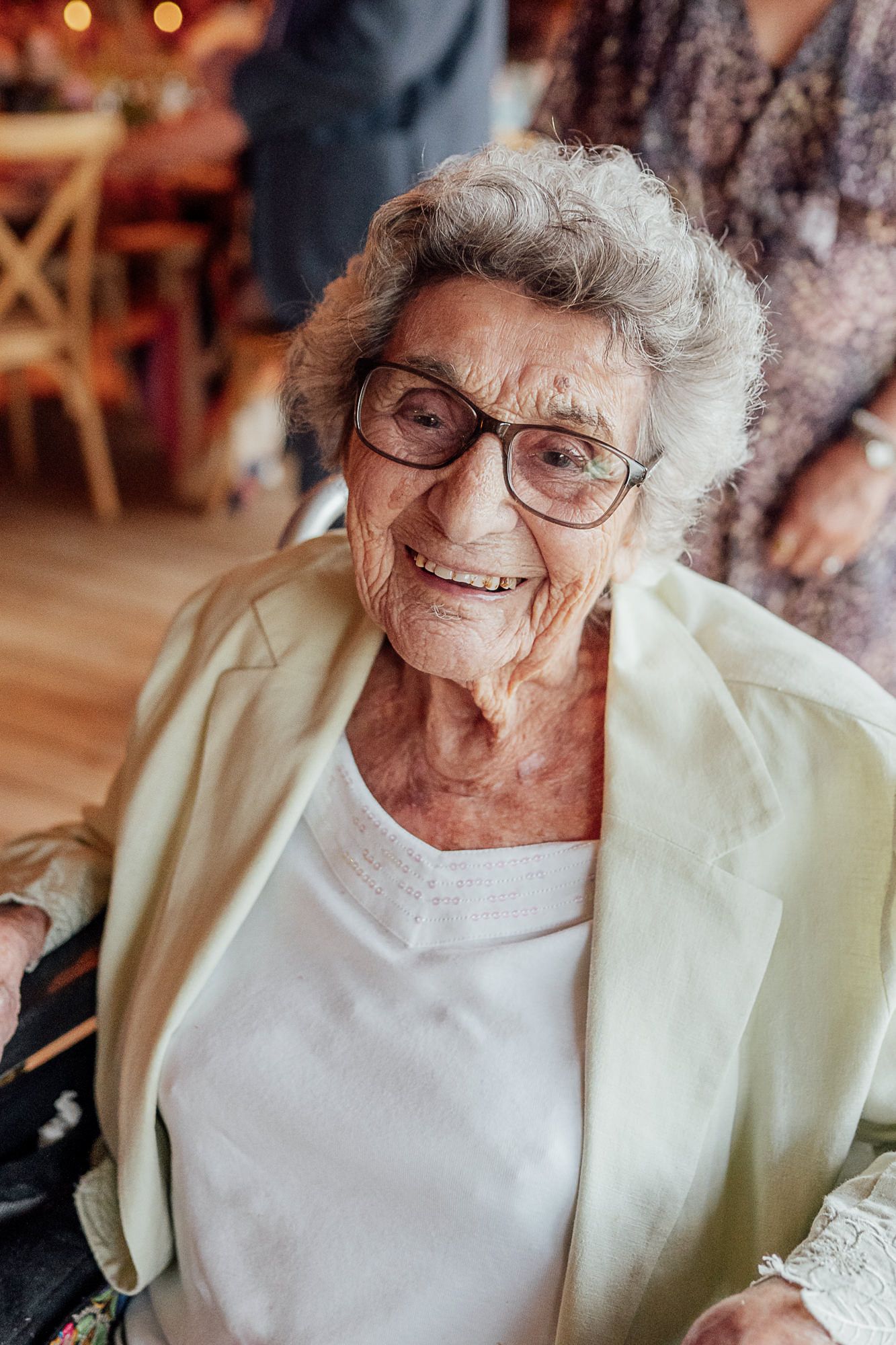 Elderly woman with gray hair and glasses smiling warmly at an indoor gathering, wearing a white top and light jacket.