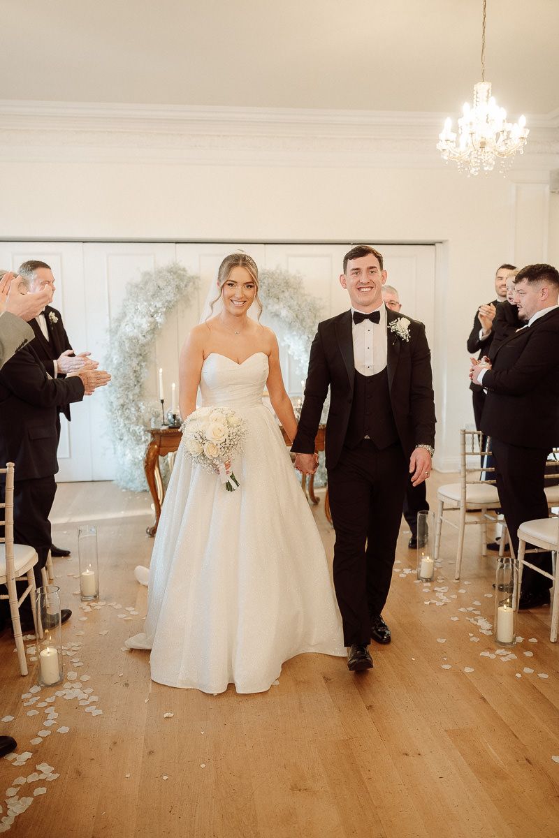 Newlyweds walk down the aisle in an elegant white room with hardwood floors and scattered rose petals.