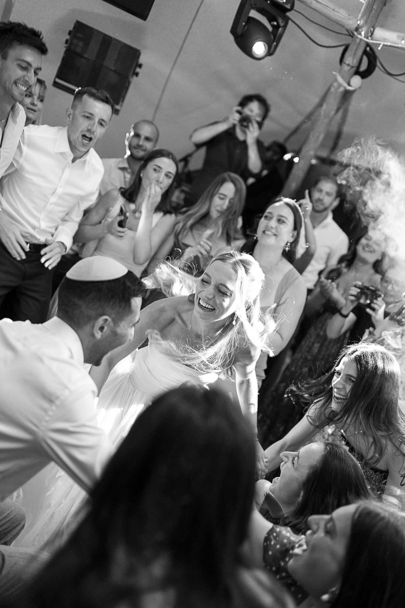 Black and white photo of a bride laughing with guests during an energetic Jewish wedding dance celebration.