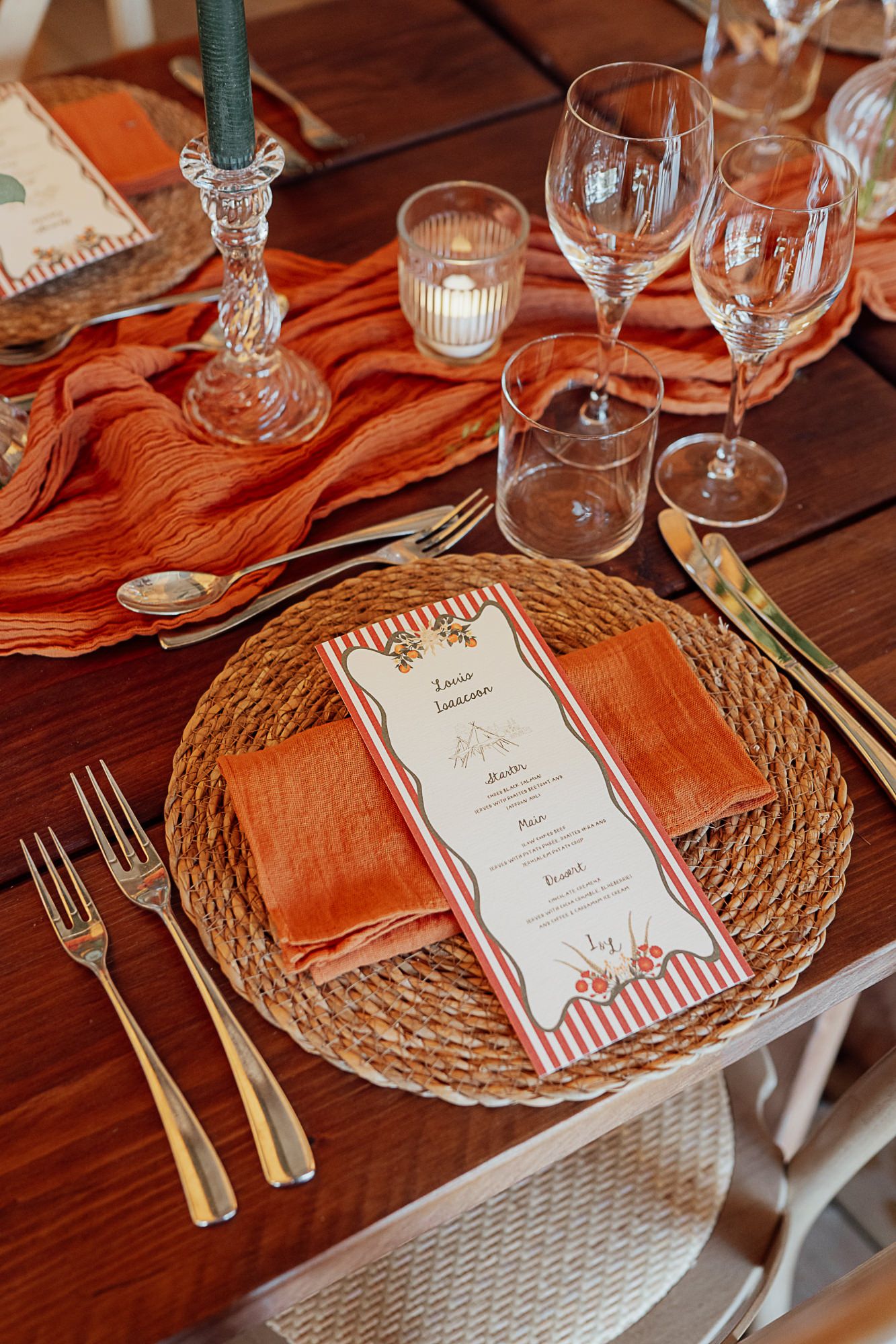 Elegant rustic wedding table setting with orange napkins, wicker charger, menu card, crystal glasses, and gold cutlery.