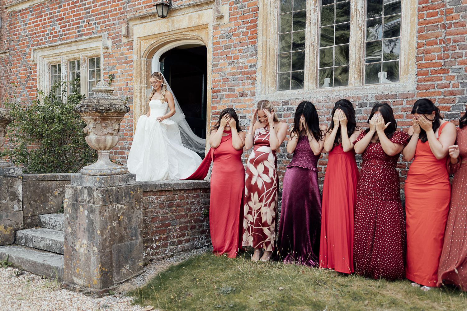 Bride in white gown stands on steps while bridesmaids in red dresses cover their faces outside a brick manor.