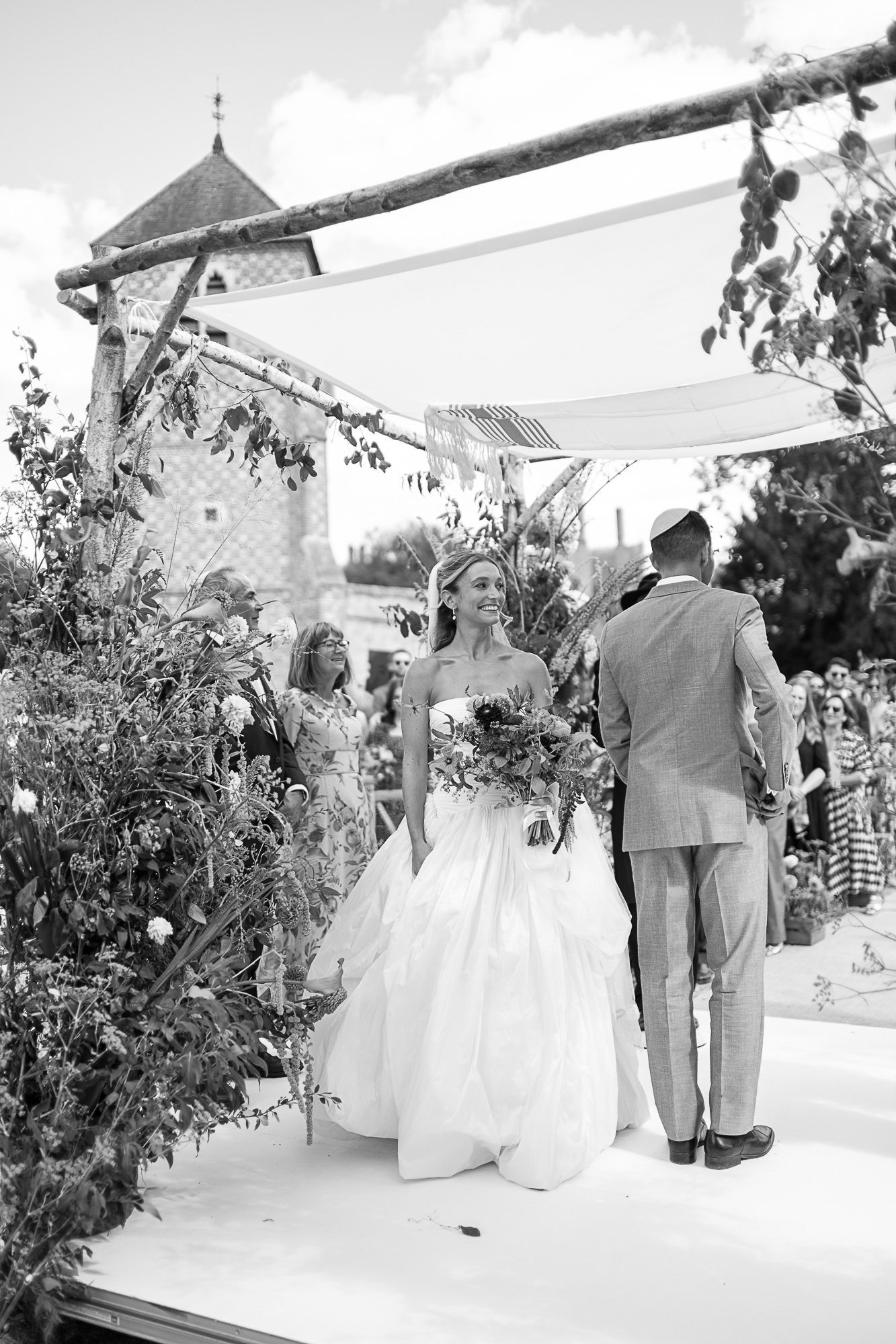 Black and white photo of a smiling bride in a white gown holding a bouquet at an outdoor wedding ceremony.