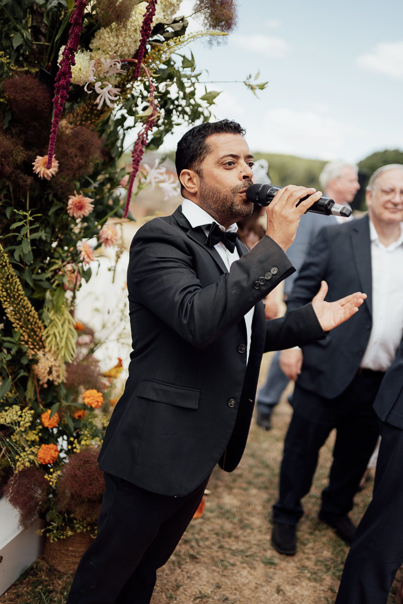 Man in black tuxedo singing into microphone at outdoor wedding ceremony with floral arch backdrop.