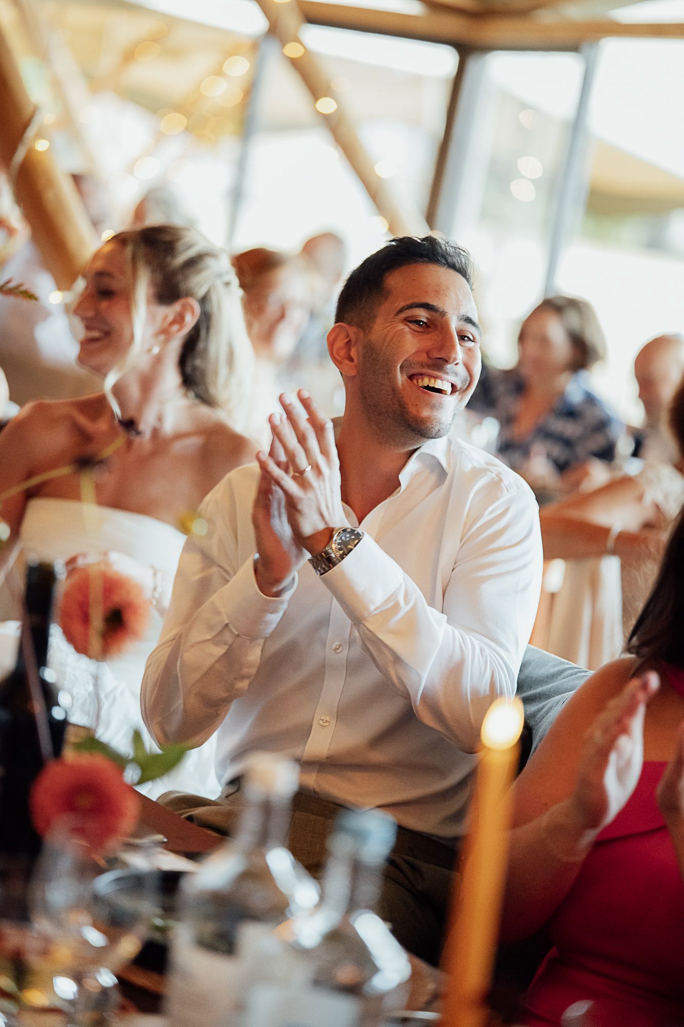 A smiling man in a white shirt claps at a wedding reception while guests celebrate around a candlelit table.