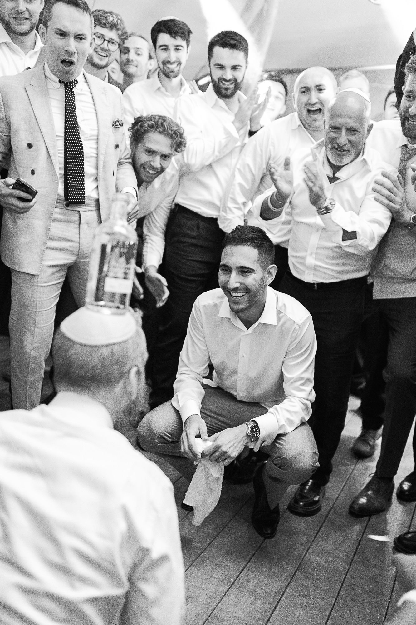 Black and white photo of men celebrating at a party, one crouching and smiling while others cheer around him.