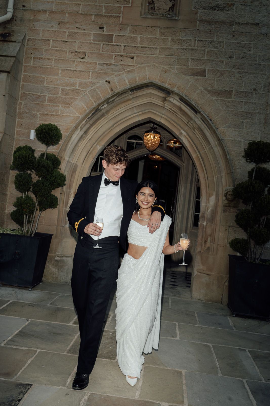 Bride in white saree and groom in black tuxedo hold champagne glasses outside stone castle venue.