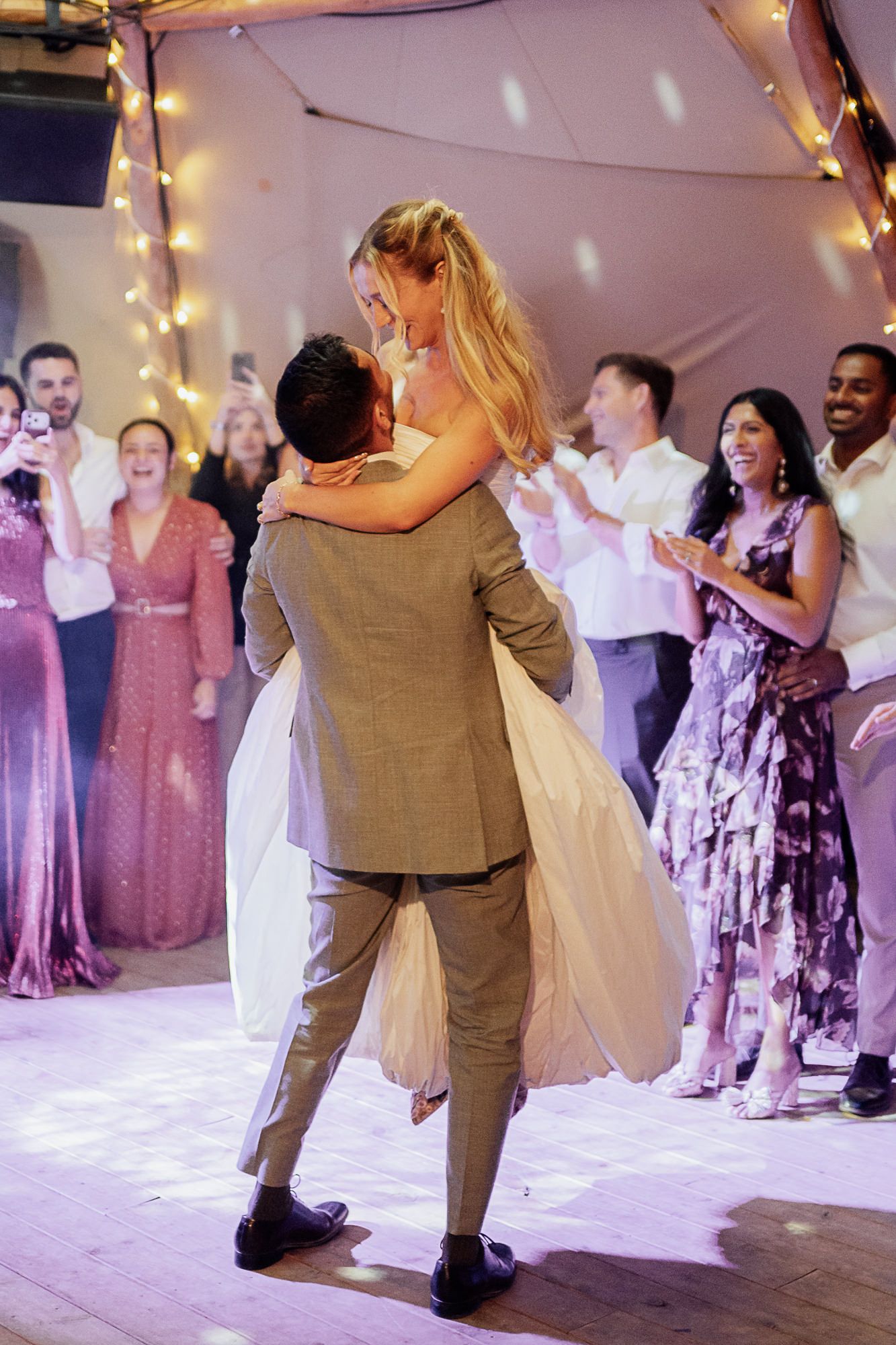 Groom lifting bride during first dance at wedding reception with smiling guests celebrating in background.