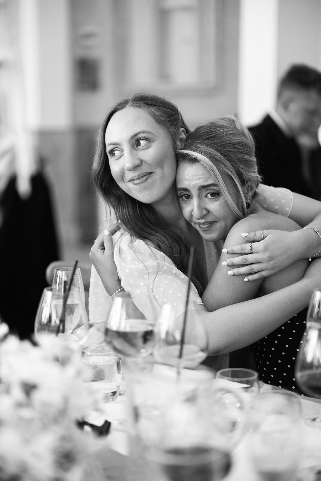 Two women share a warm hug at a wedding reception table in black and white photo.