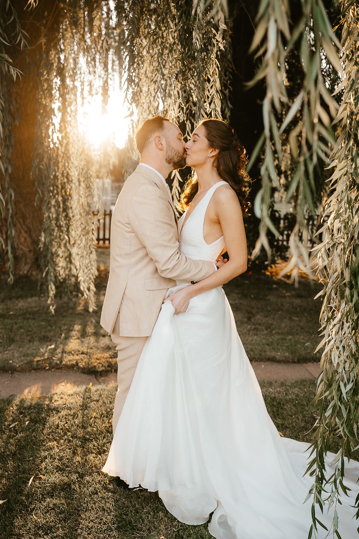 Romantic sunset wedding portrait under willow trees with golden light filtering through branches.
