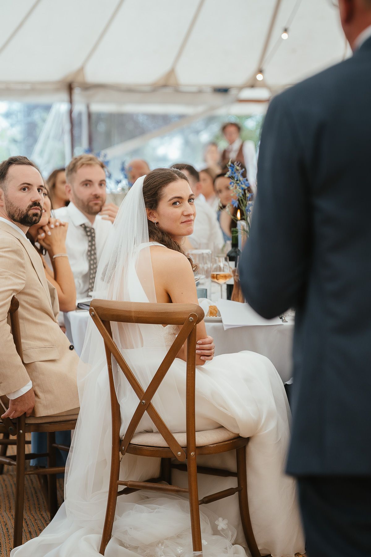 Guests at a wedding reception under a white tent with wooden cross-back chairs and elegant decor.