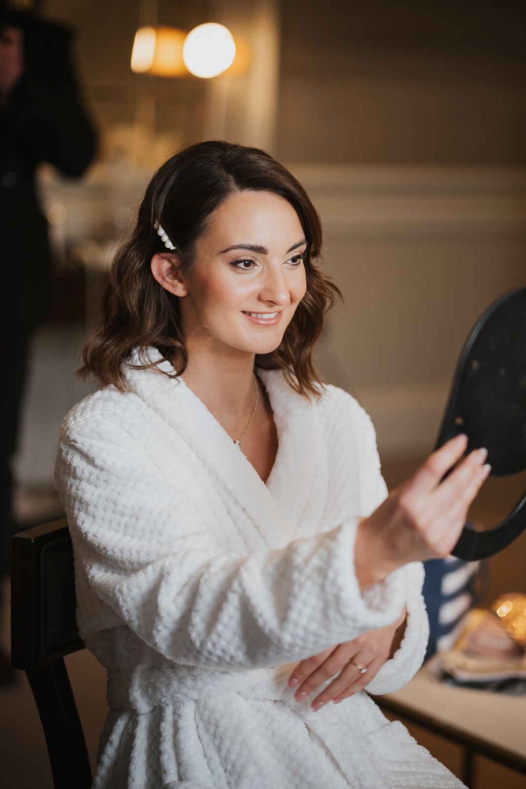 A woman in a white bathrobe smiles while looking at herself in a mirror during preparation.