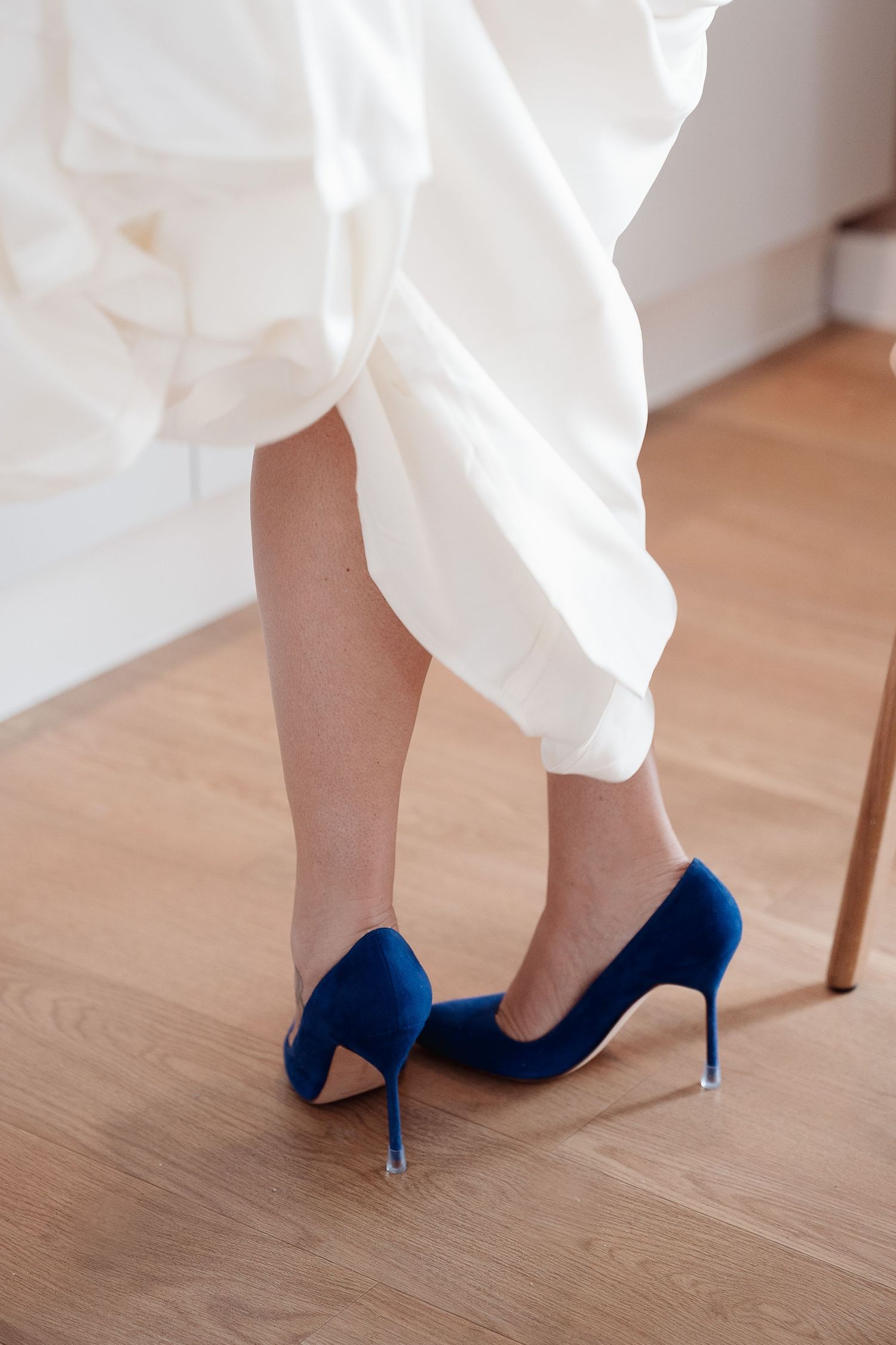 Elegant royal blue velvet high heel pumps worn with white pants on a hardwood floor.