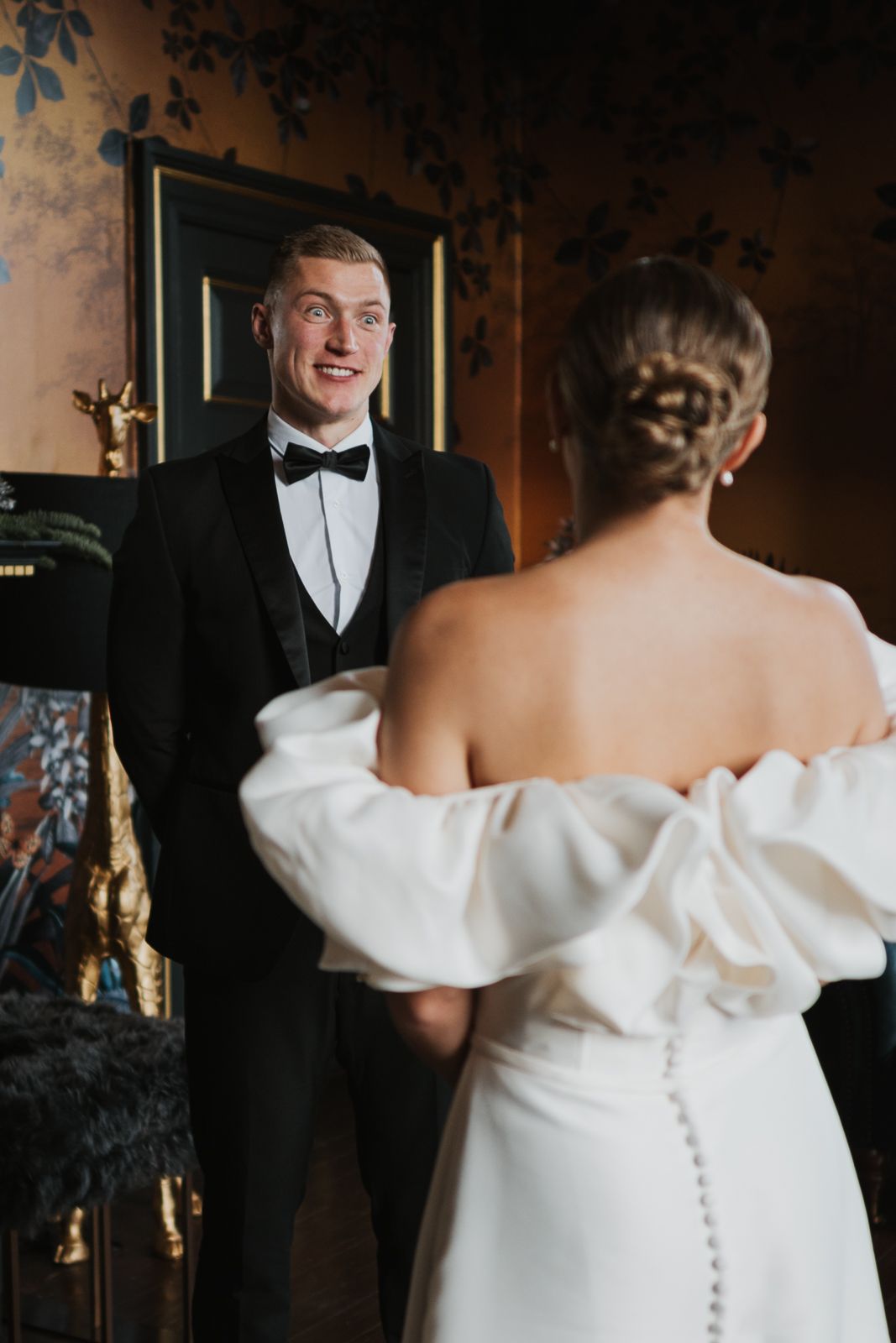 A romantic first look moment between a bride in an off-shoulder wedding dress and groom in classic black tuxedo.