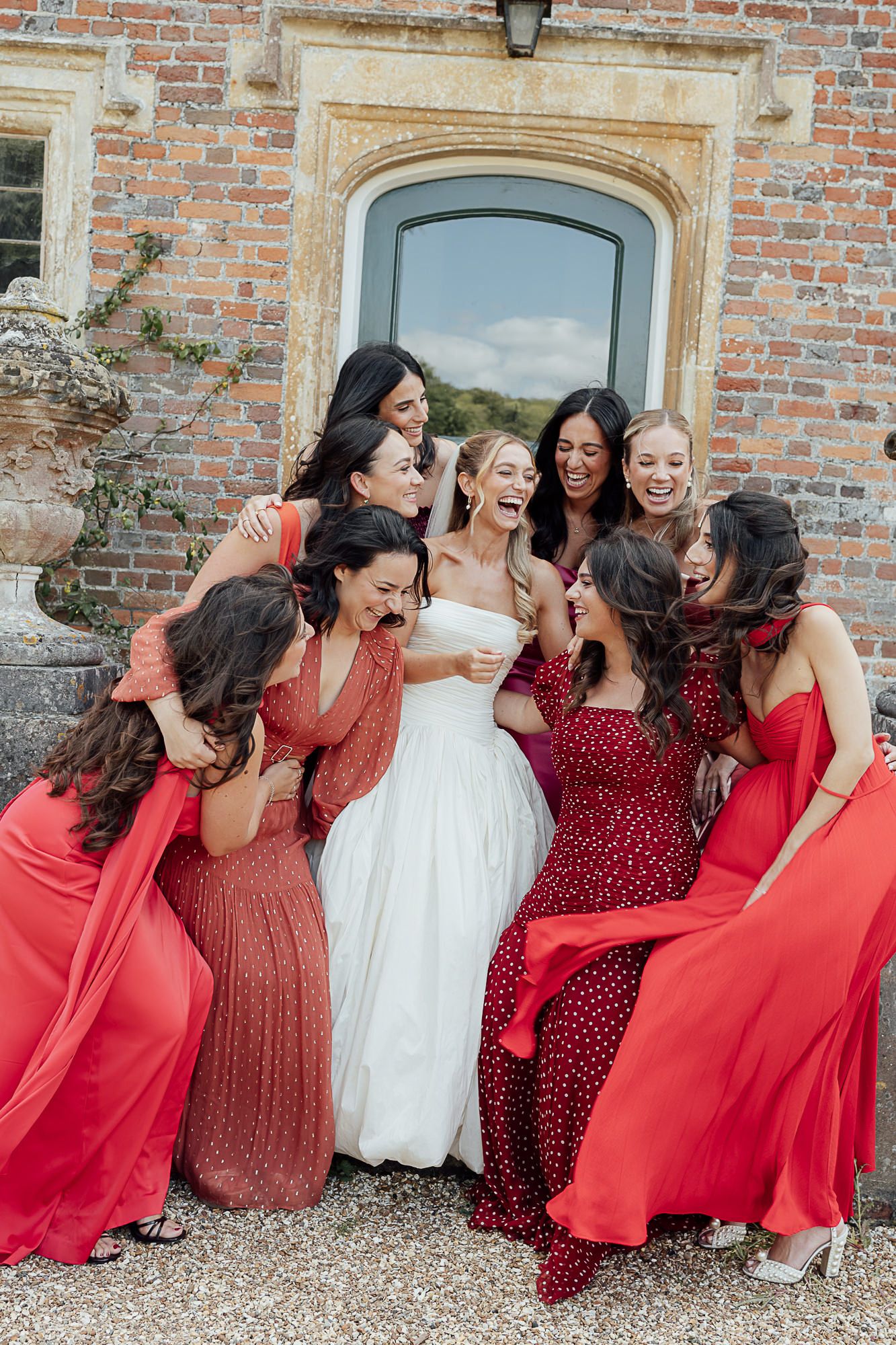 Bride in white gown laughing with bridesmaids in red dresses outside a brick manor house.