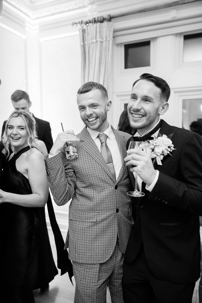 A group of groomsmen in black suits and one in a checkered vest share candid laughs during a wedding celebration.