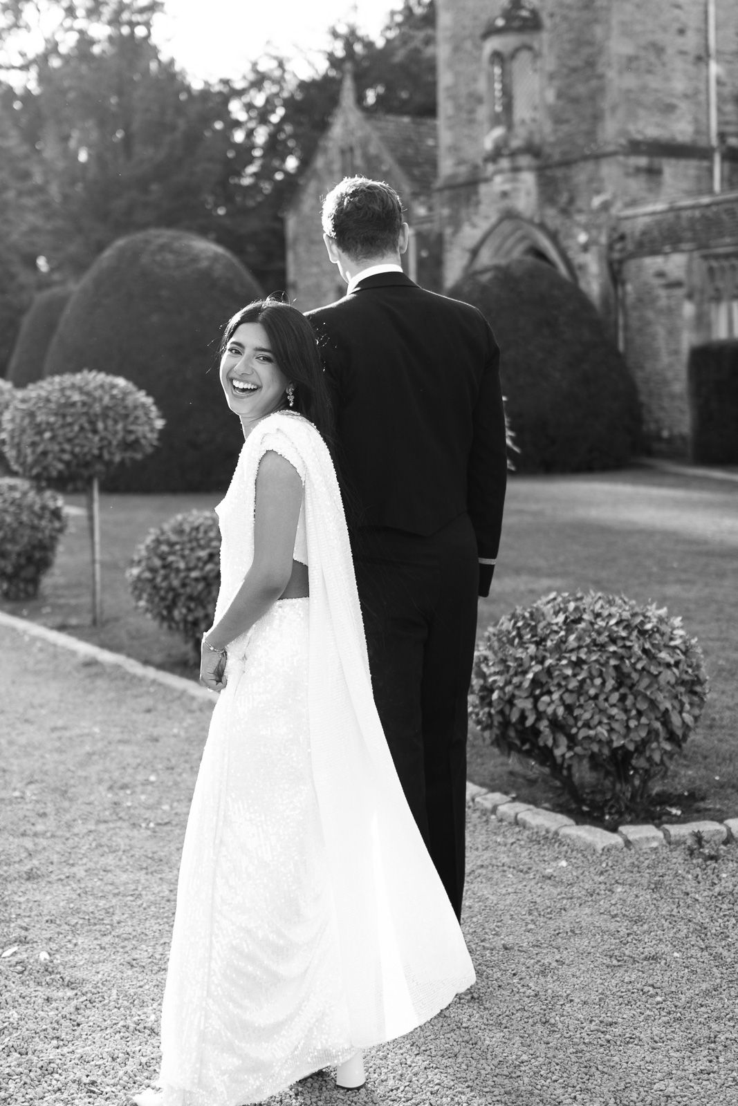 Bride in white sari laughs over shoulder while groom in tuxedo walks through elegant garden.