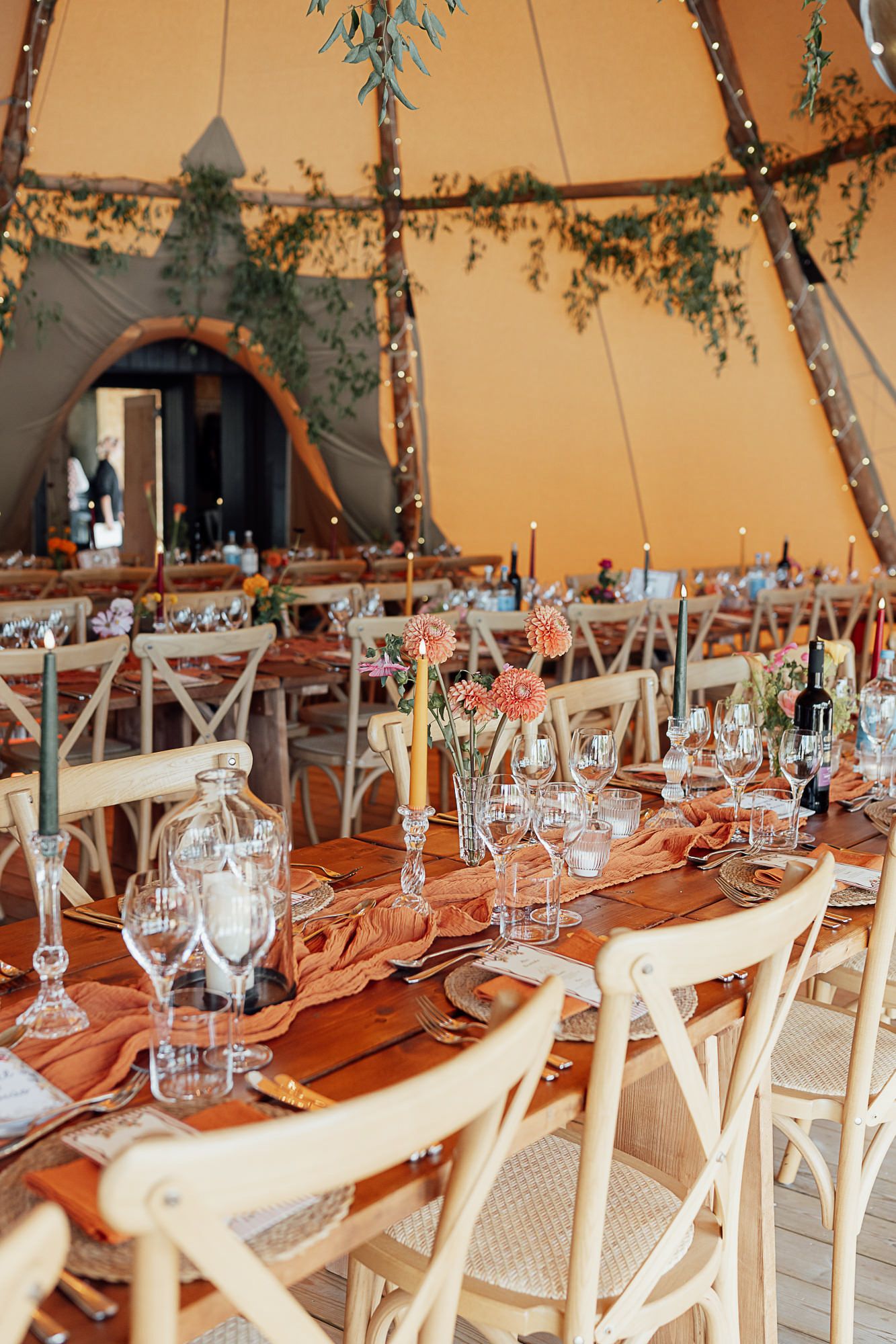 Elegant tipi wedding reception with rustic wooden chairs, floral centerpieces, candles, and terracotta table runners.