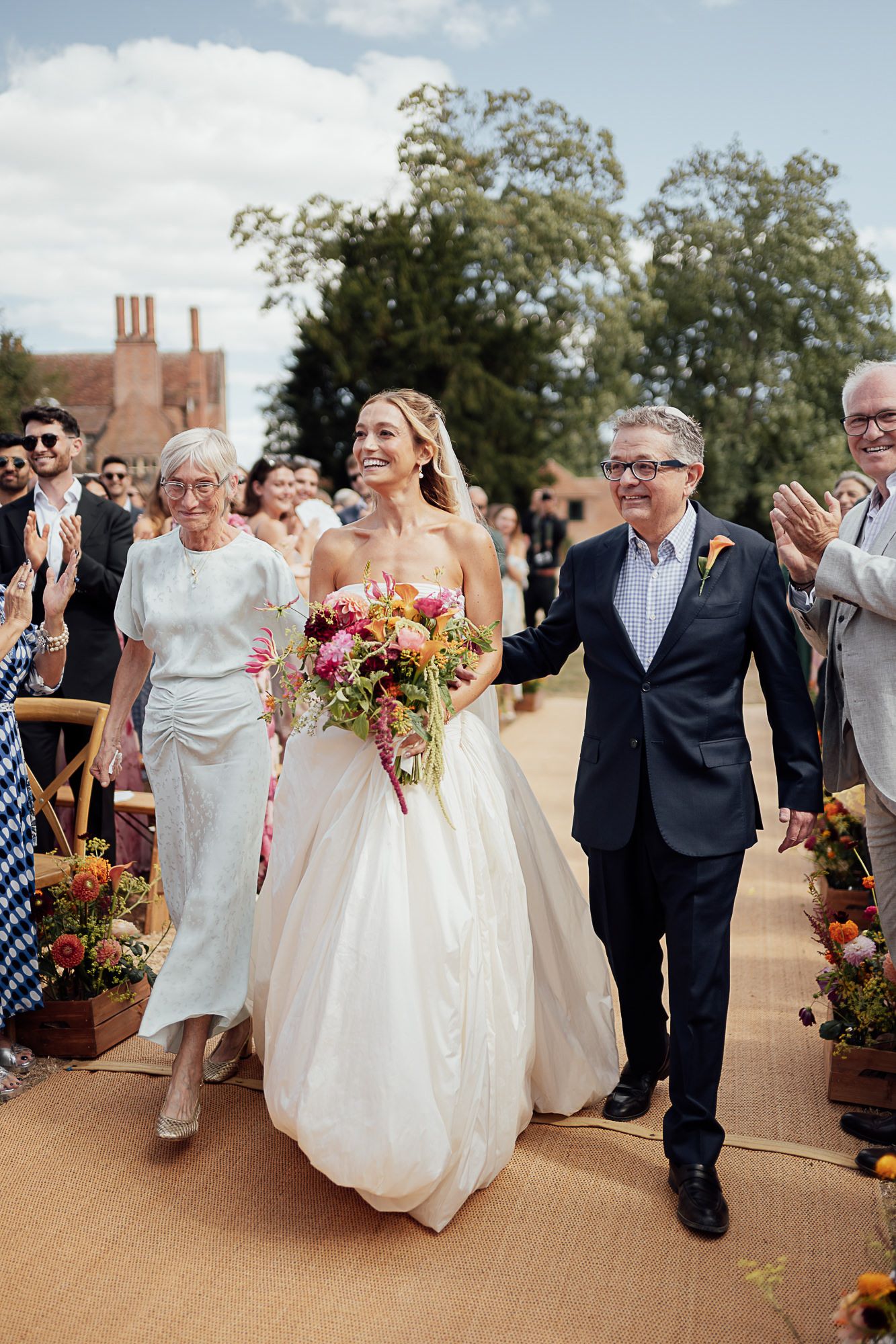 Bride walks down outdoor aisle with family, holding vibrant bouquet, guests applauding, lush greenery backdrop.