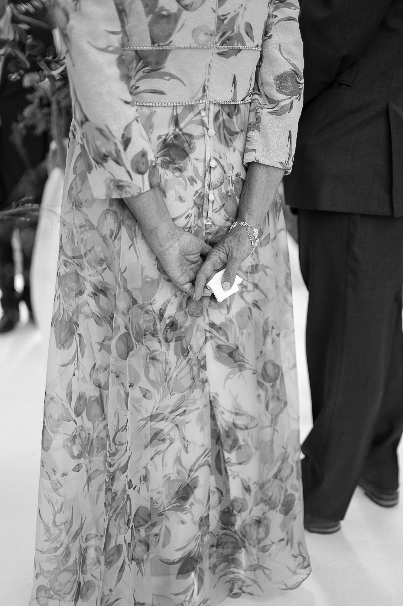 Black and white photo of elderly woman in floral dress clasping hands, holding tissue at formal event.