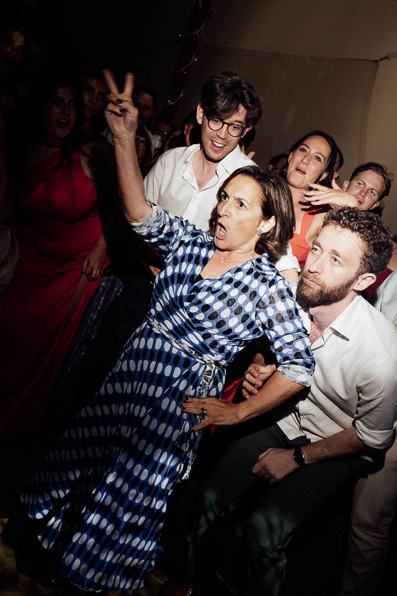Excited party guests dancing and posing, woman in blue patterned dress making peace sign at lively indoor celebration.