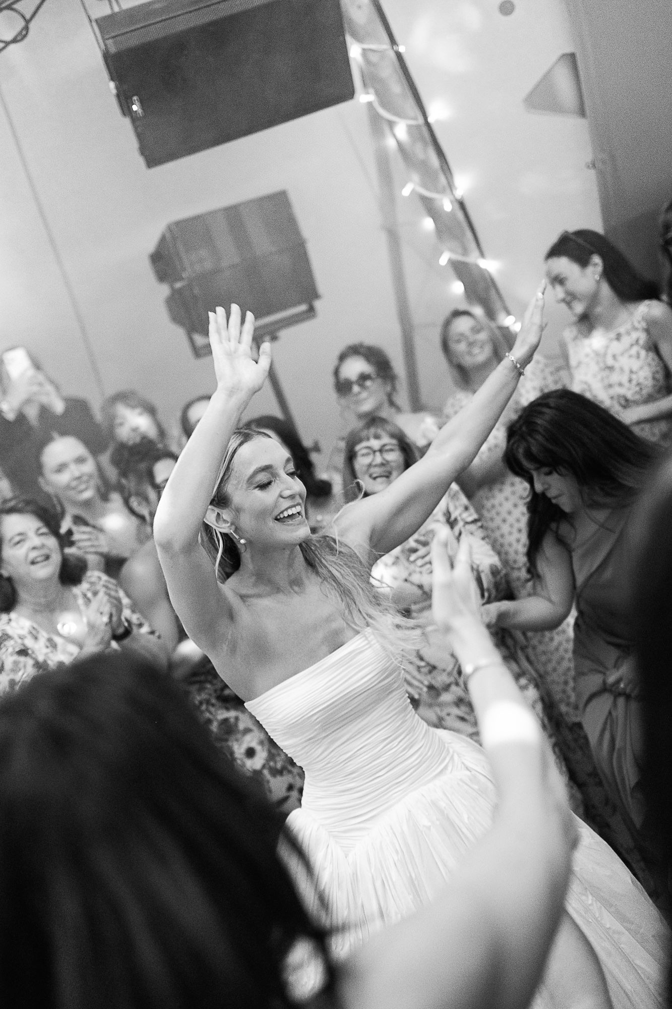 Bride in white strapless gown celebrates joyfully with raised arms surrounded by smiling wedding guests in black and white photo.