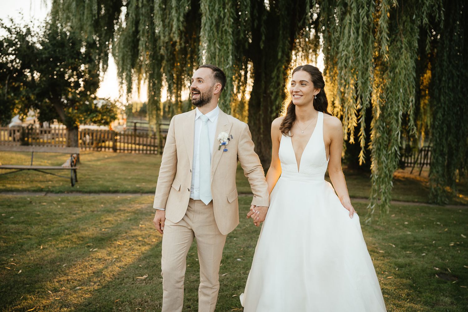 A couple in wedding attire stands together beneath a willow tree during sunset in a rustic outdoor setting.