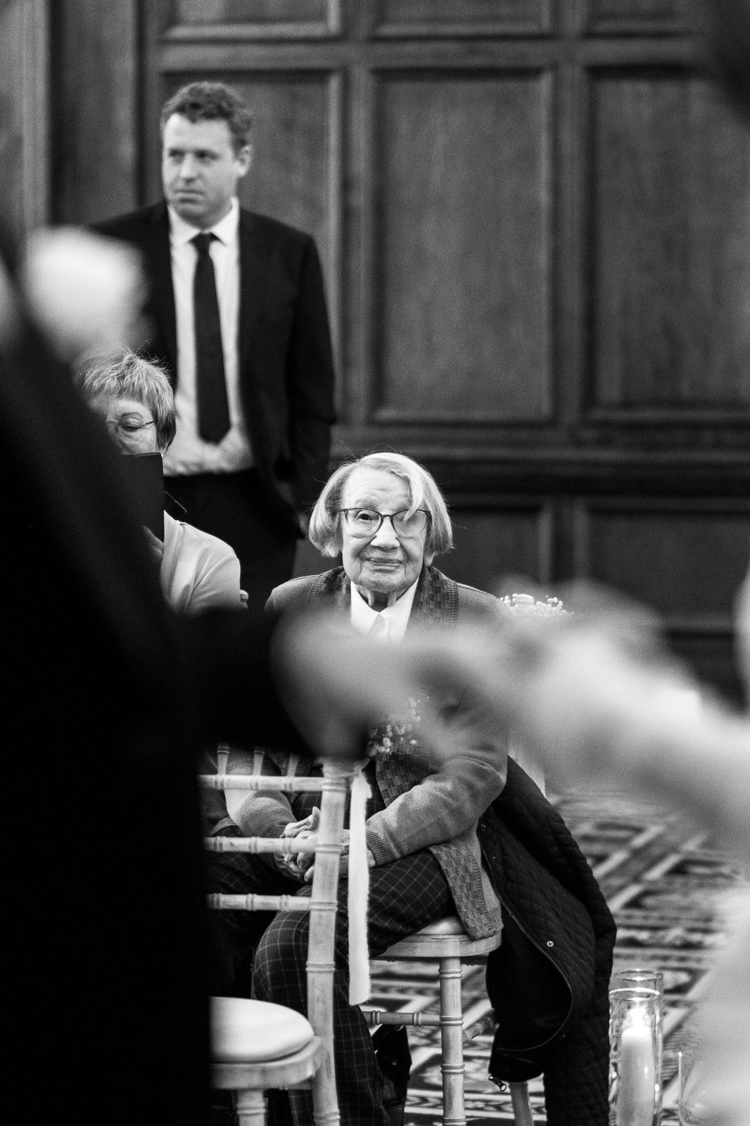 Black and white candid photo of guests seated in formal wood-paneled venue during ceremony.