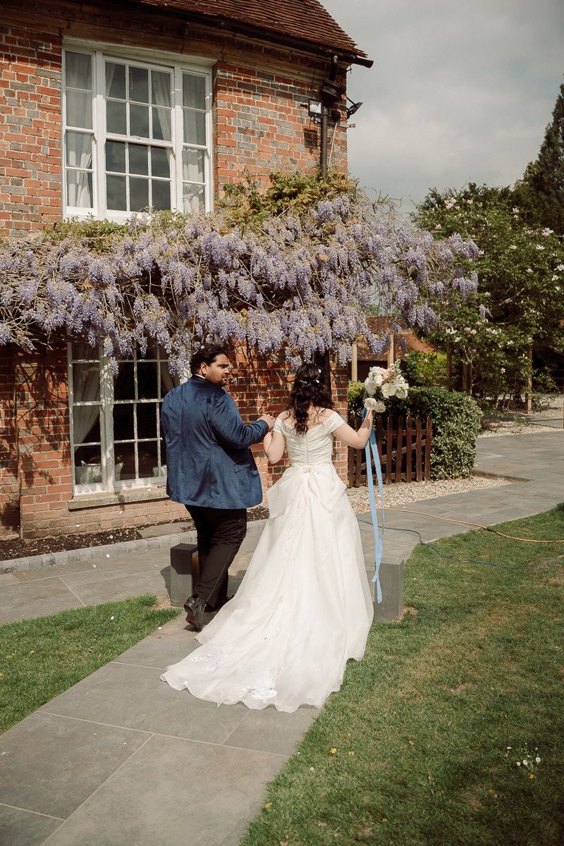 A couple walks along a brick building exterior with blooming wisteria flowers cascading from above.