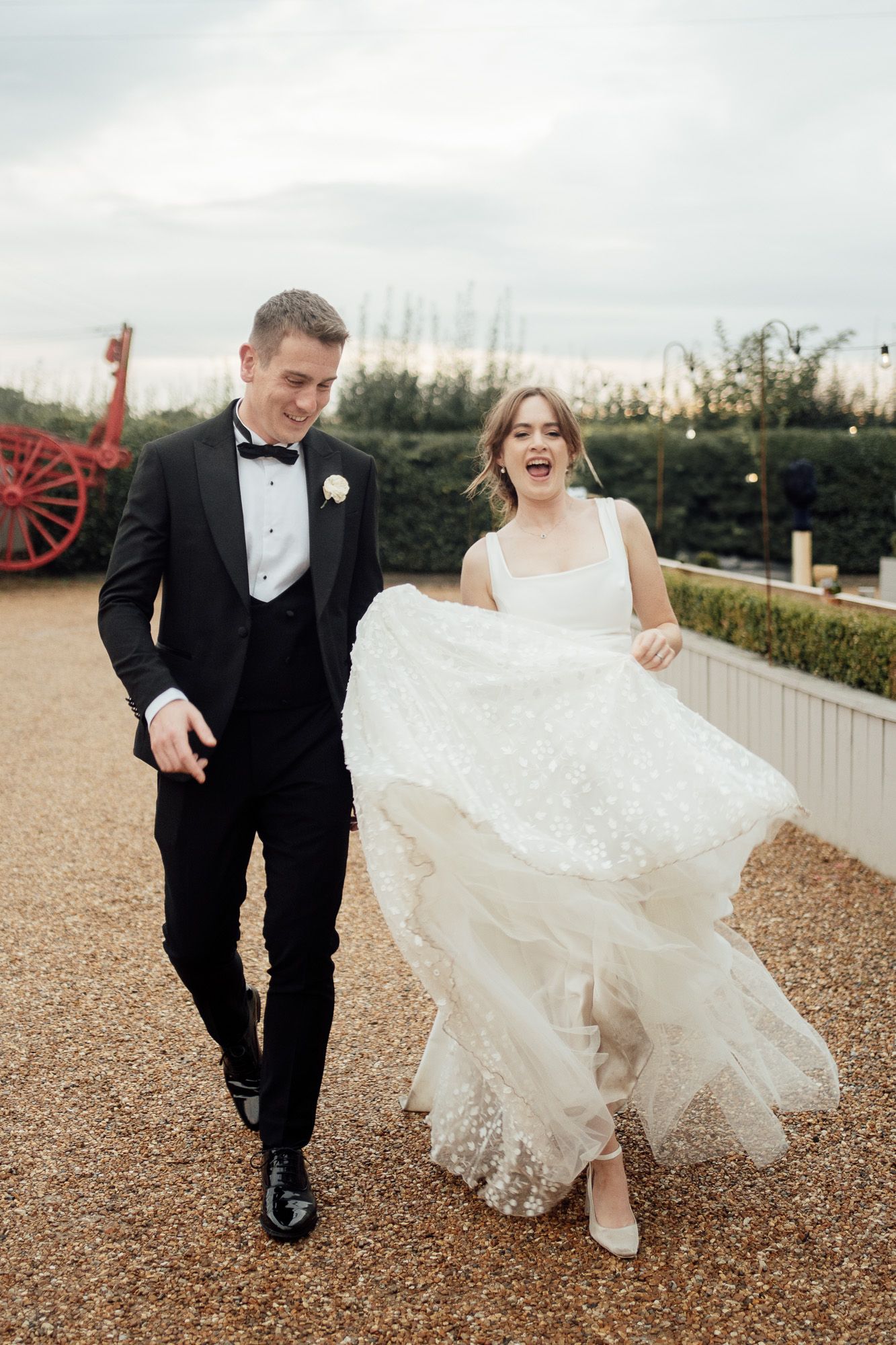 A newlywed couple walking together on a gravel path, with the bride's white lace dress flowing in the breeze.